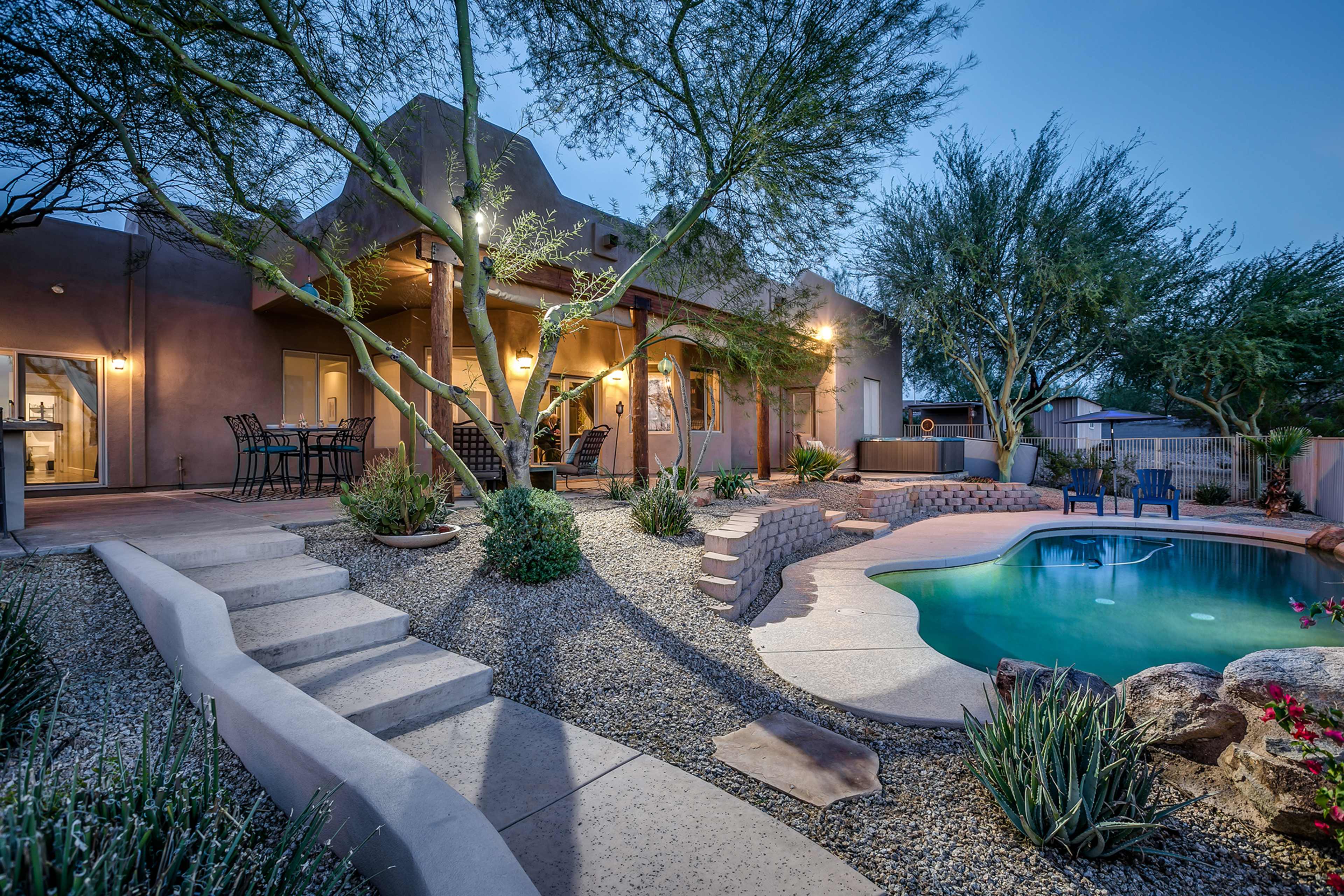 A home with a backyard featuring a swimming pool, landscaped gardens, and patio seating under tree branches at dusk.