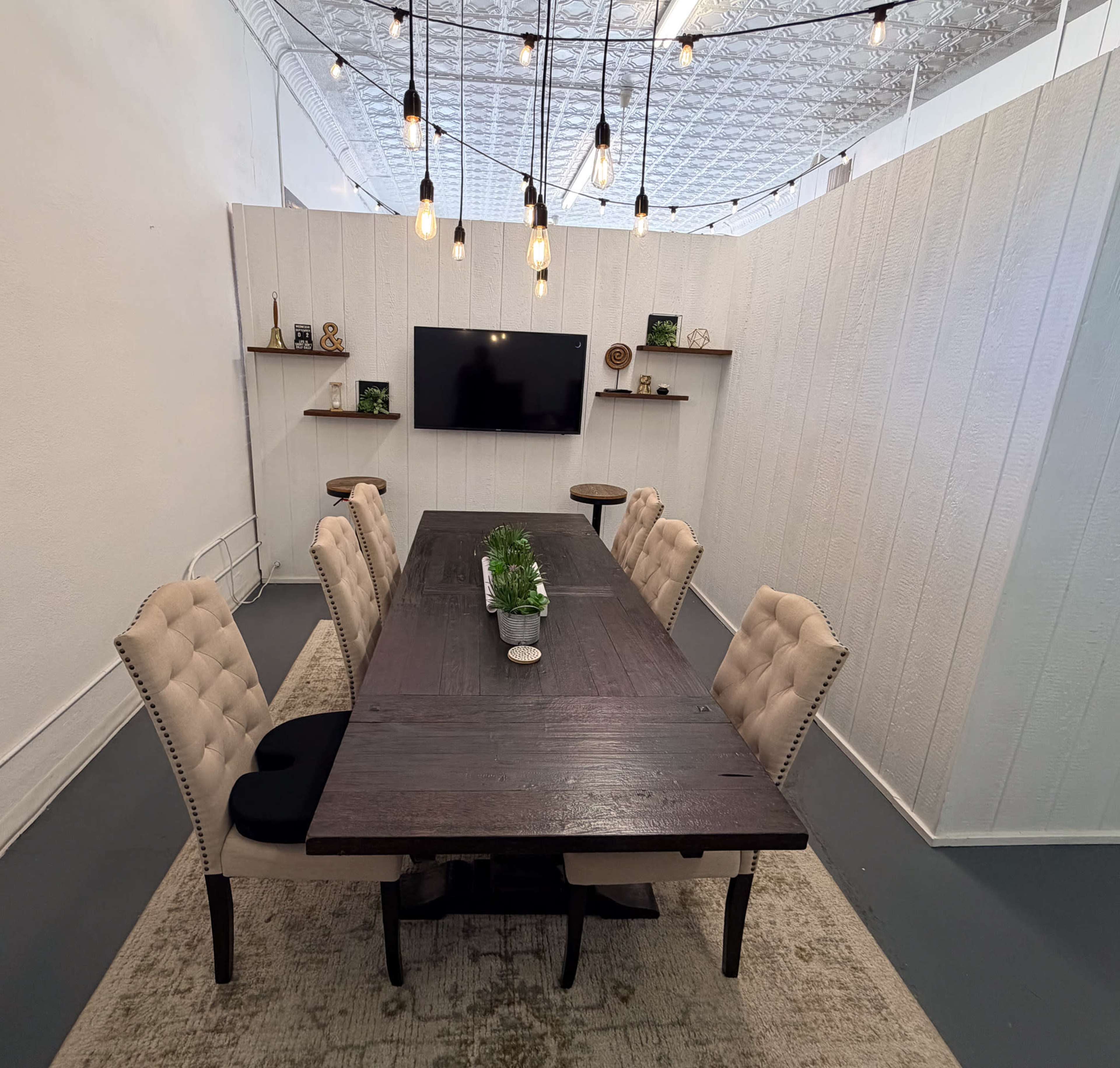 A modern meeting room features a large wooden table surrounded by upholstered chairs, with wall-mounted shelves, a television, and decorative plants.