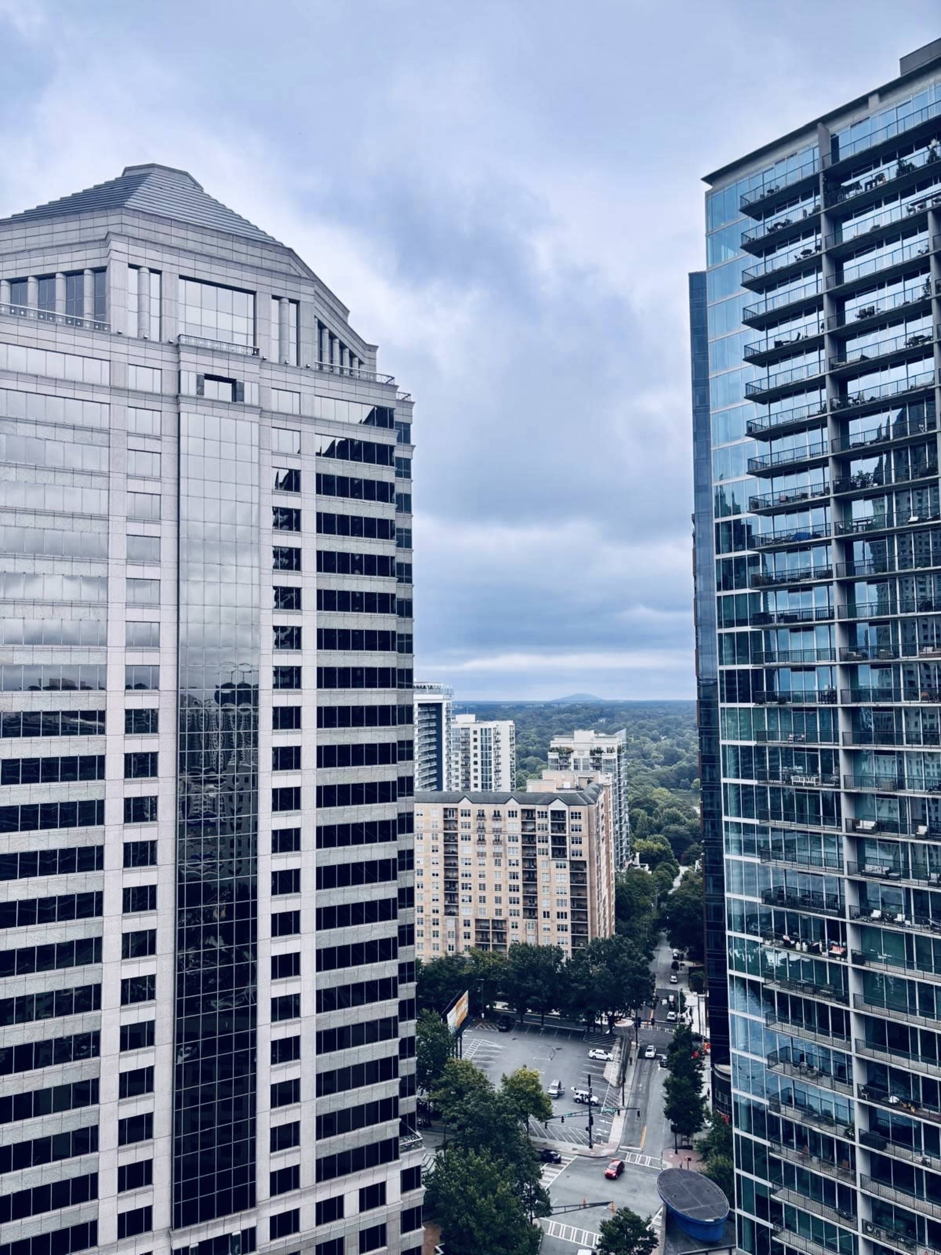 The image shows a cityscape featuring tall glass skyscrapers on either side, with mid-rise buildings and tree-lined streets visible in the distance under a cloudy sky.