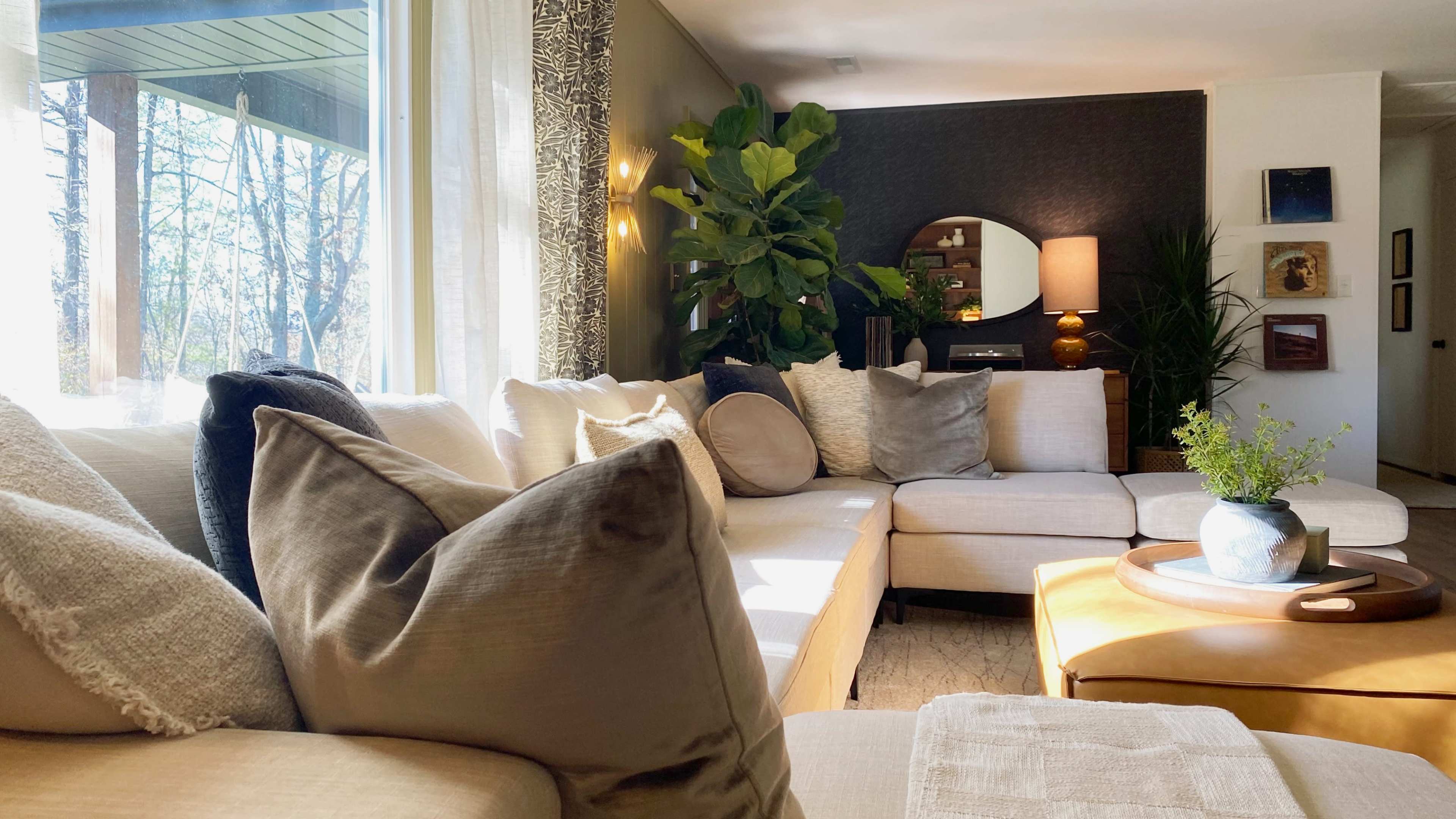 The image shows a modern living room featuring a sectional sofa with assorted pillows, a round mirror, and indoor plants, illuminated by natural light from large windows.