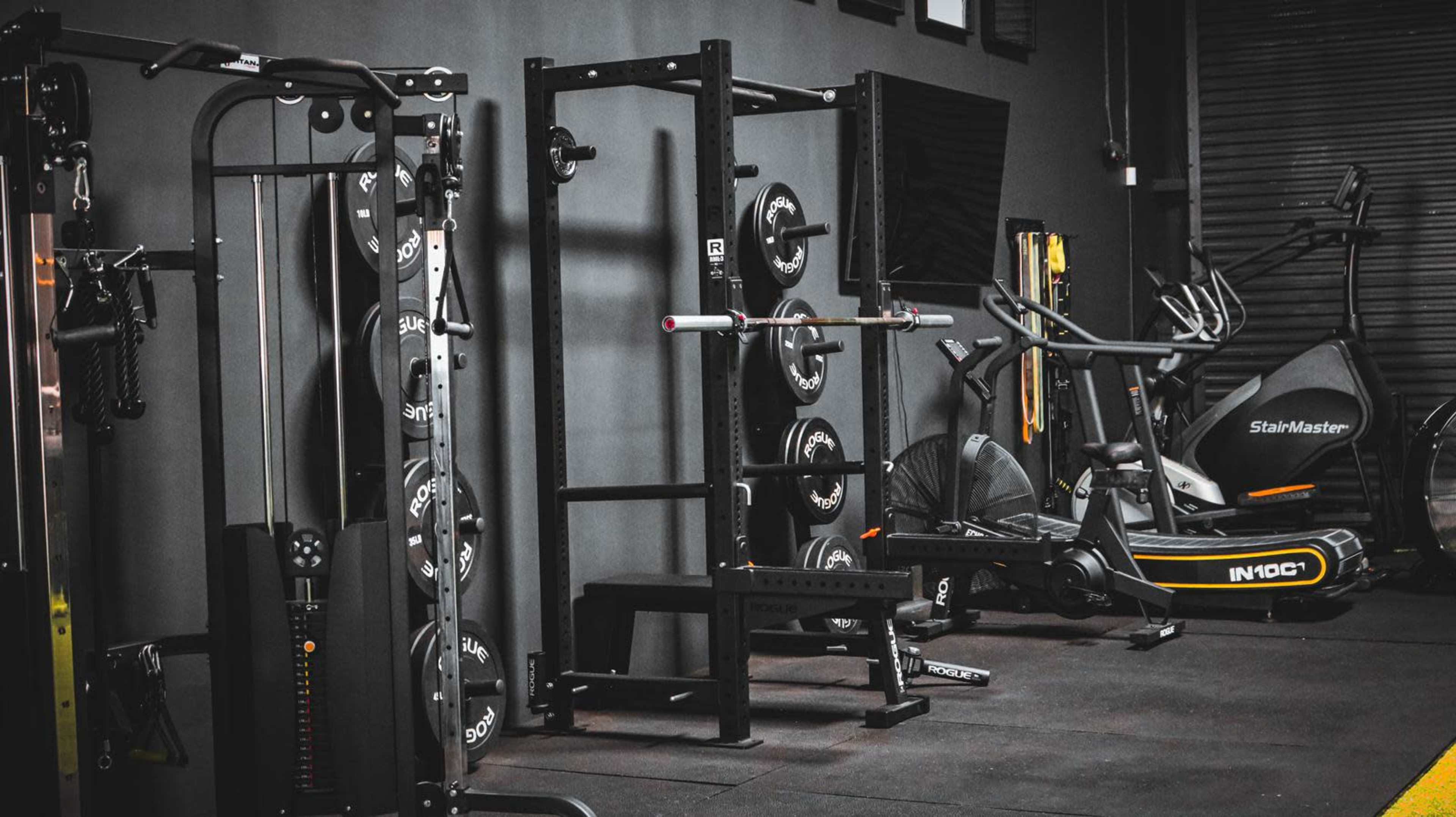 A gym interior featuring various exercise equipment including a squat rack, weight plates, and an elliptical machine.