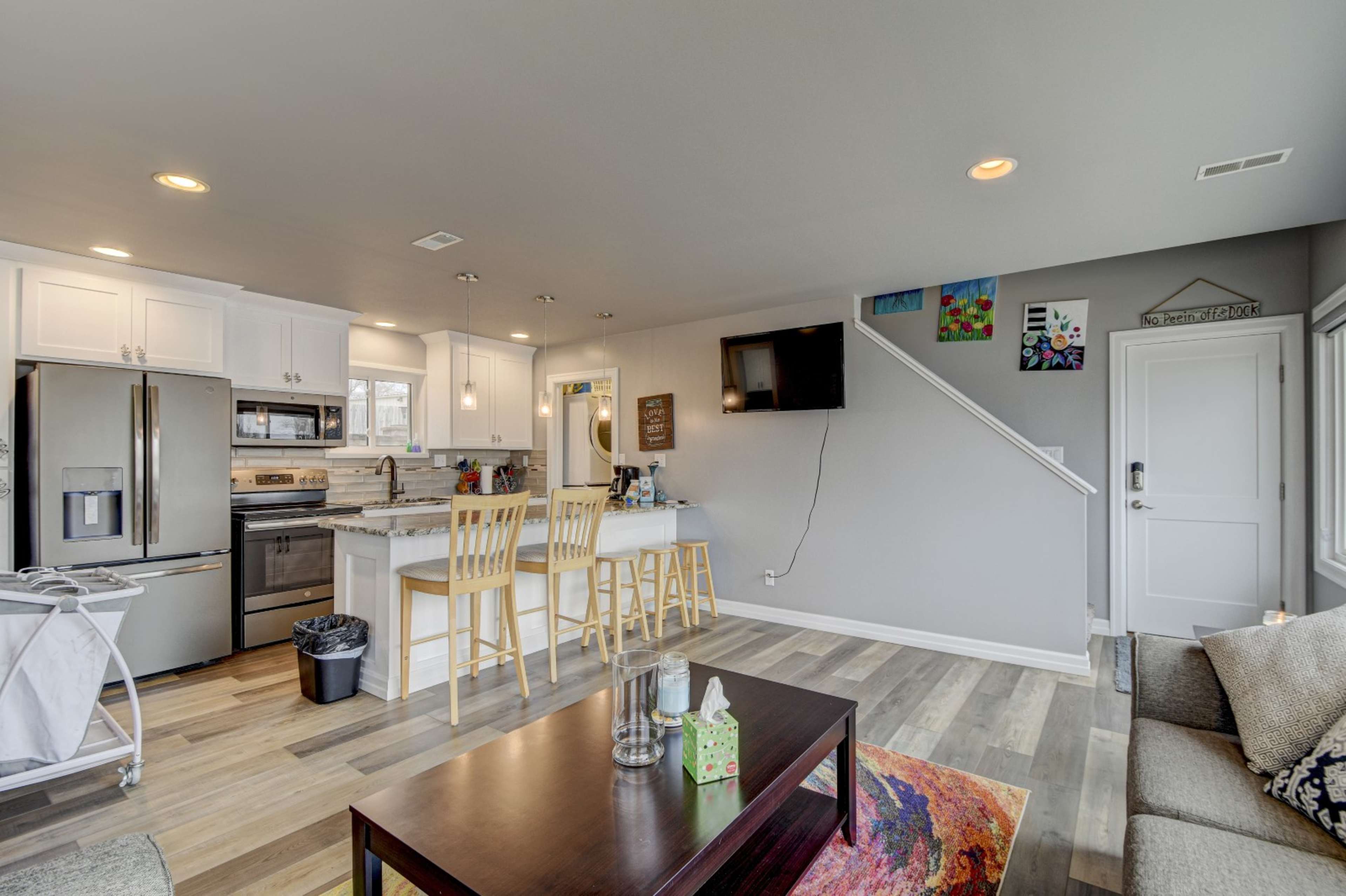 A modern open-concept living area with a kitchen featuring white cabinets, a dining table with two chairs, a sitting area with a sofa, and a television mounted on the wall.