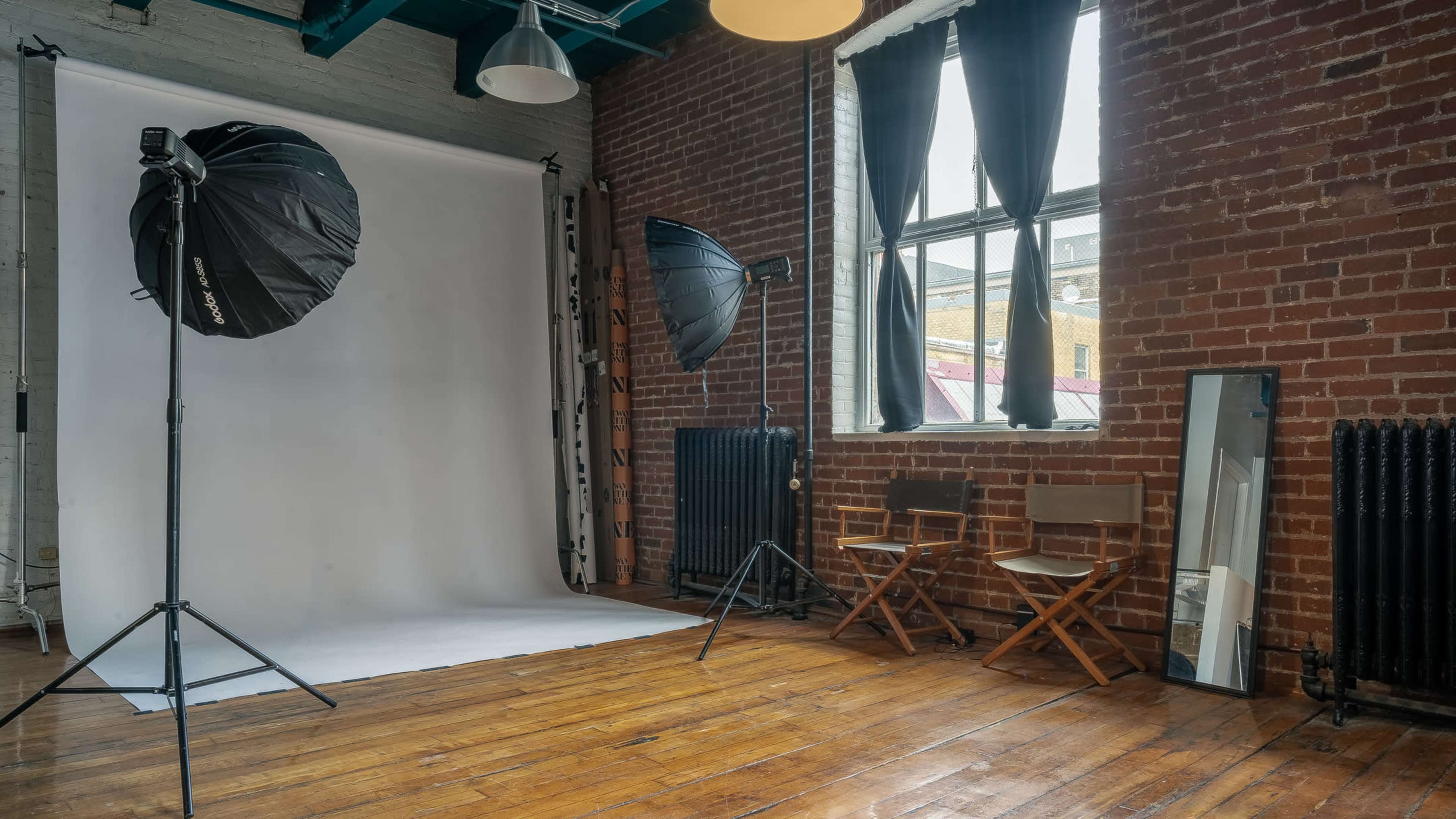 A photography studio features two large softbox lights, a backdrop, and two chairs positioned near a window with industrial-style elements.