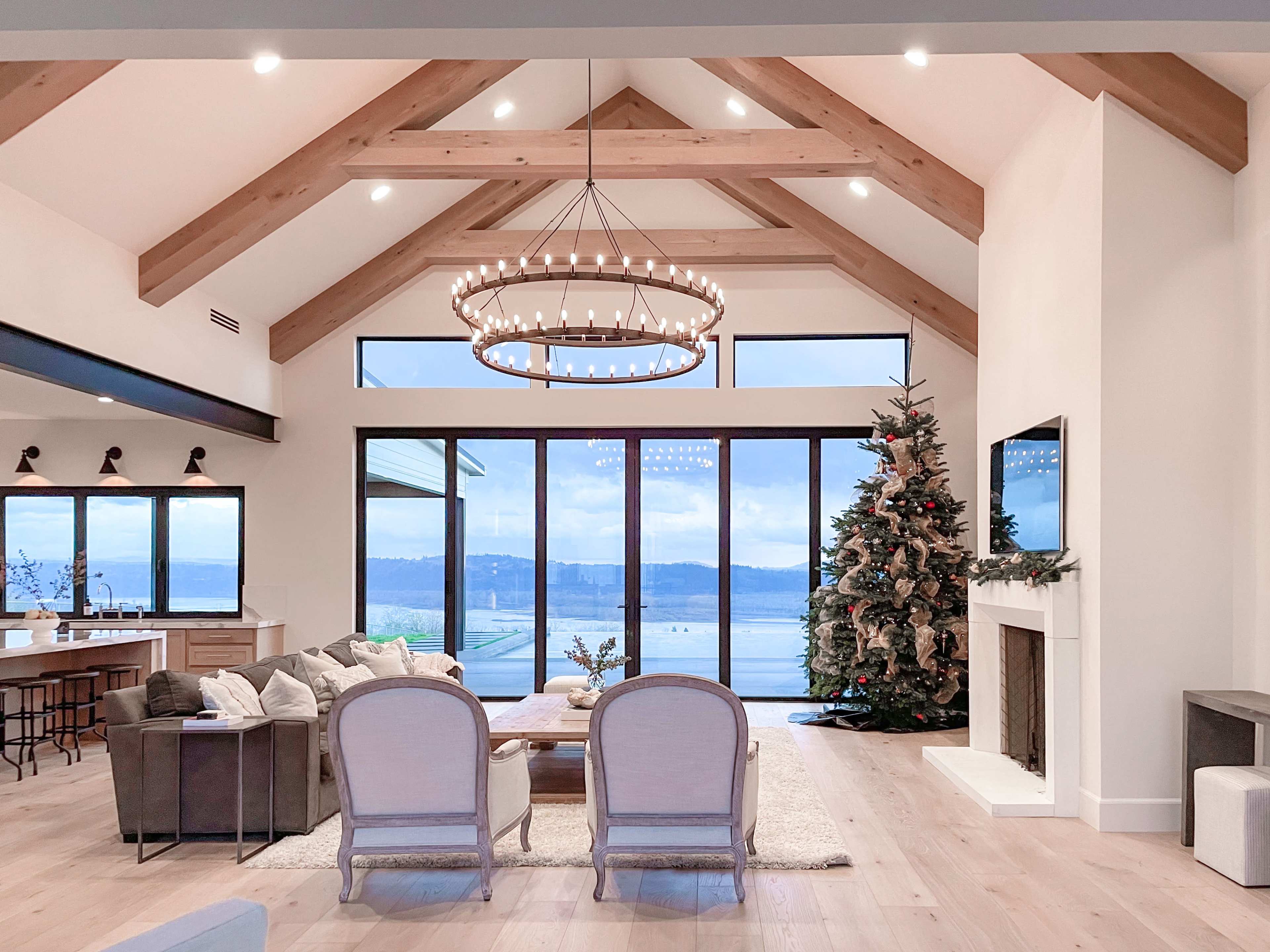 A modern living room features large windows with a view of the water, a Christmas tree in the corner, and a chandelier hanging from the vaulted ceiling.
