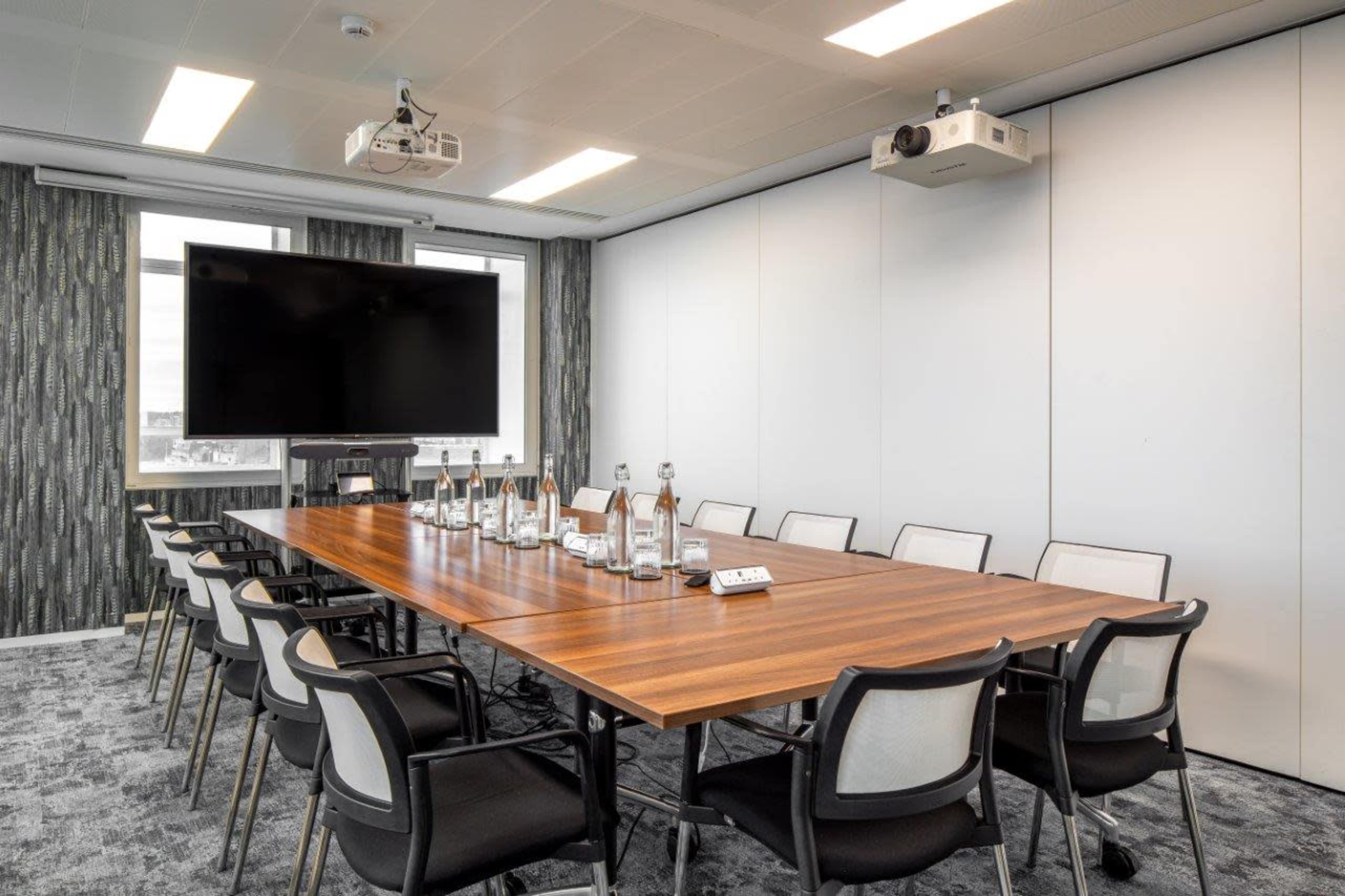 A modern conference room features a long wooden table surrounded by chairs, with a large screen and projector mounted on the wall.