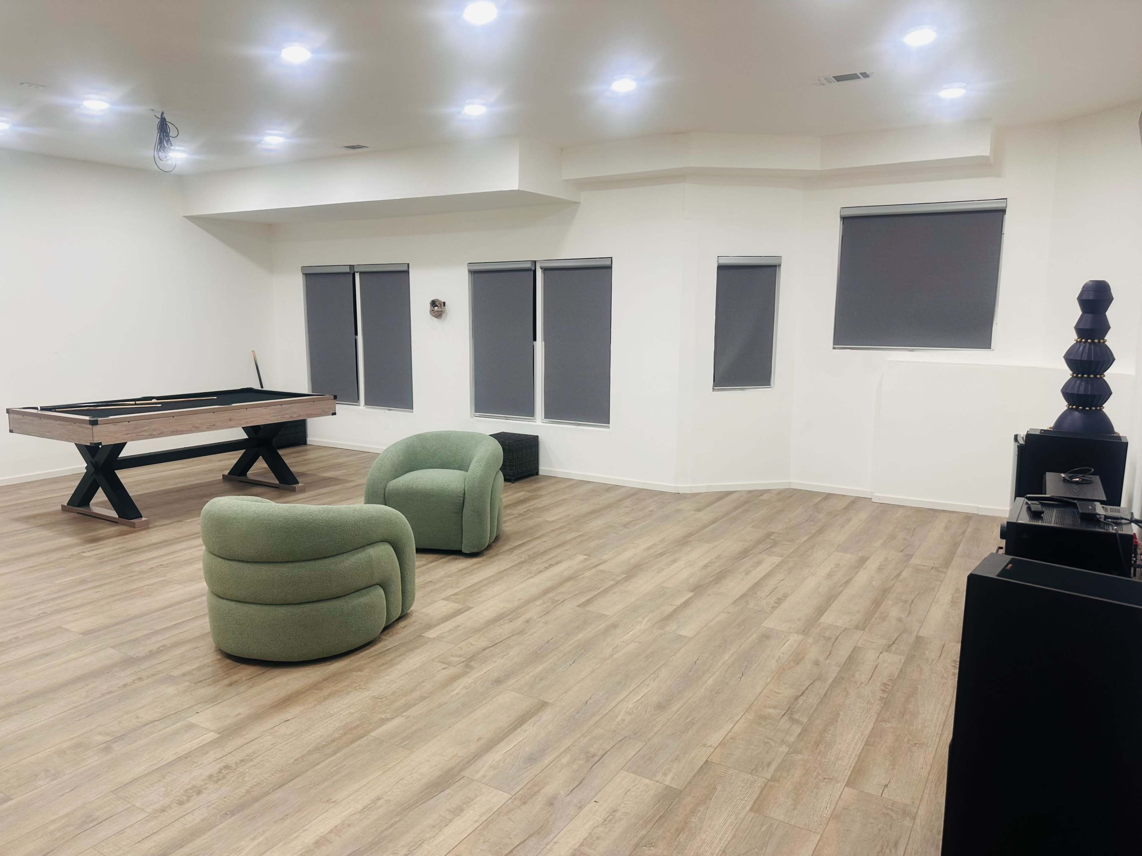 The image shows a spacious, modern room with a pool table, two green chairs, and large windows covered by gray blinds.