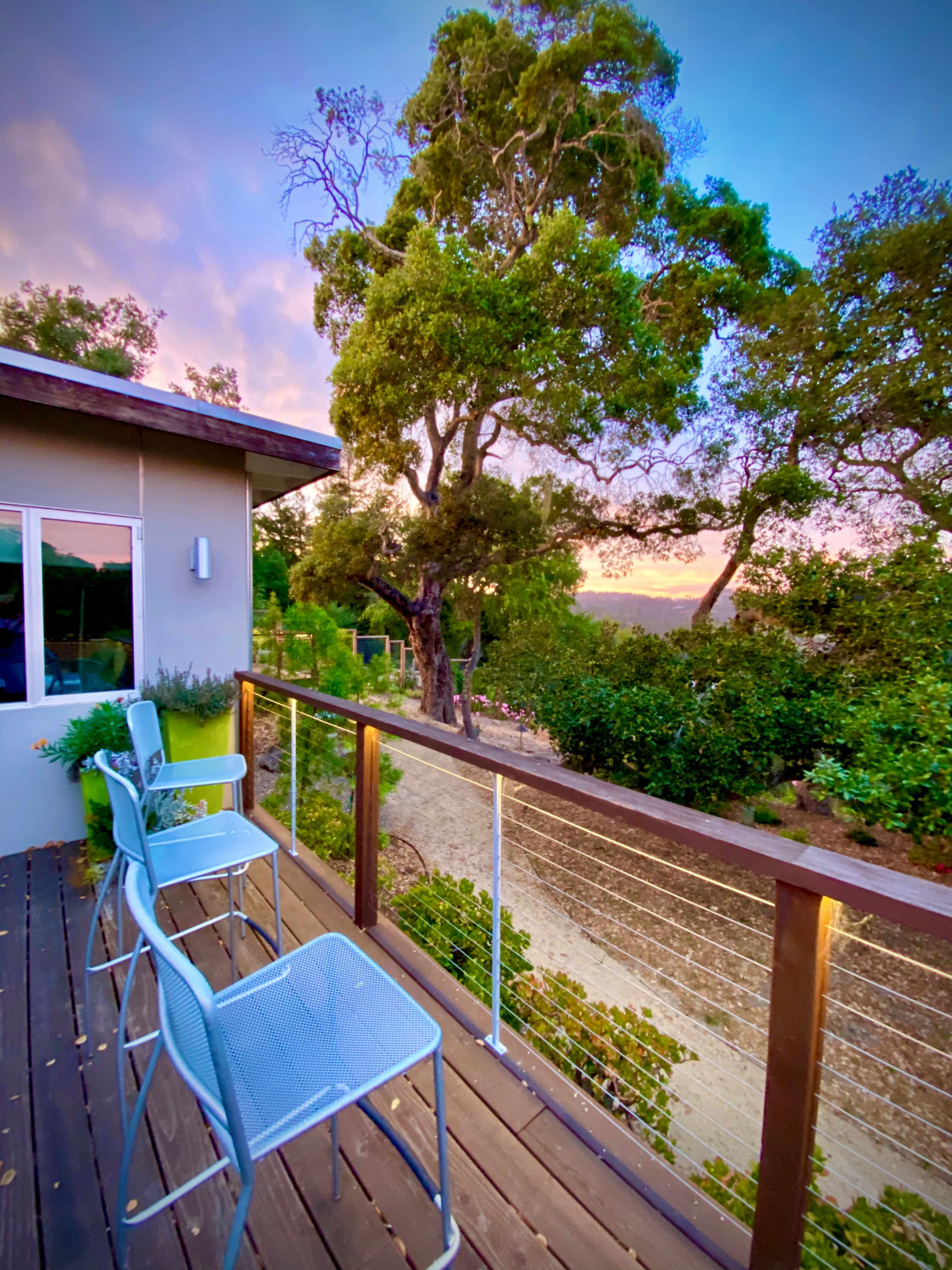 A modern balcony features three metal chairs overlooking a landscaped area with trees at sunset.
