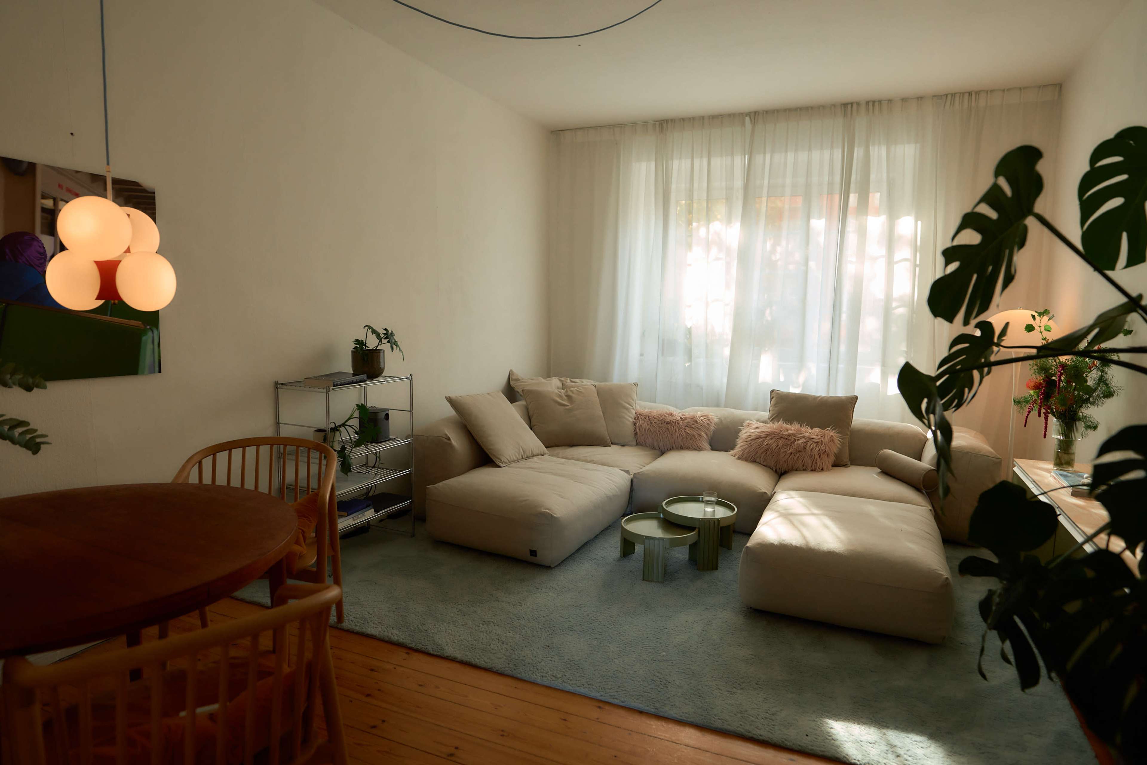 The living room features a large sectional sofa, a round dining table, and plants, with natural light filtering through sheer curtains.