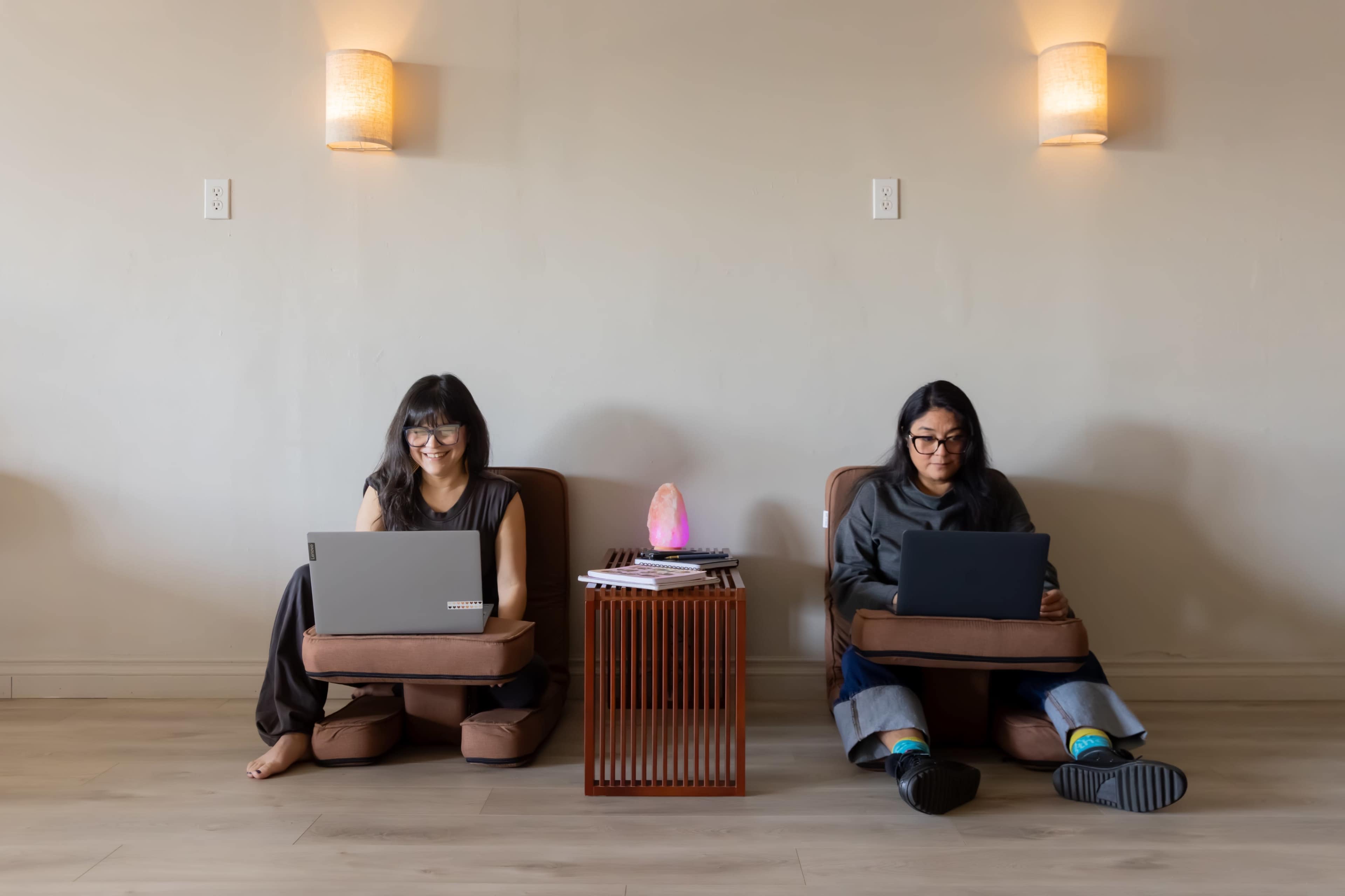 Two people sit on floor cushions with laptops in a softly lit room, separated by a small wooden table.