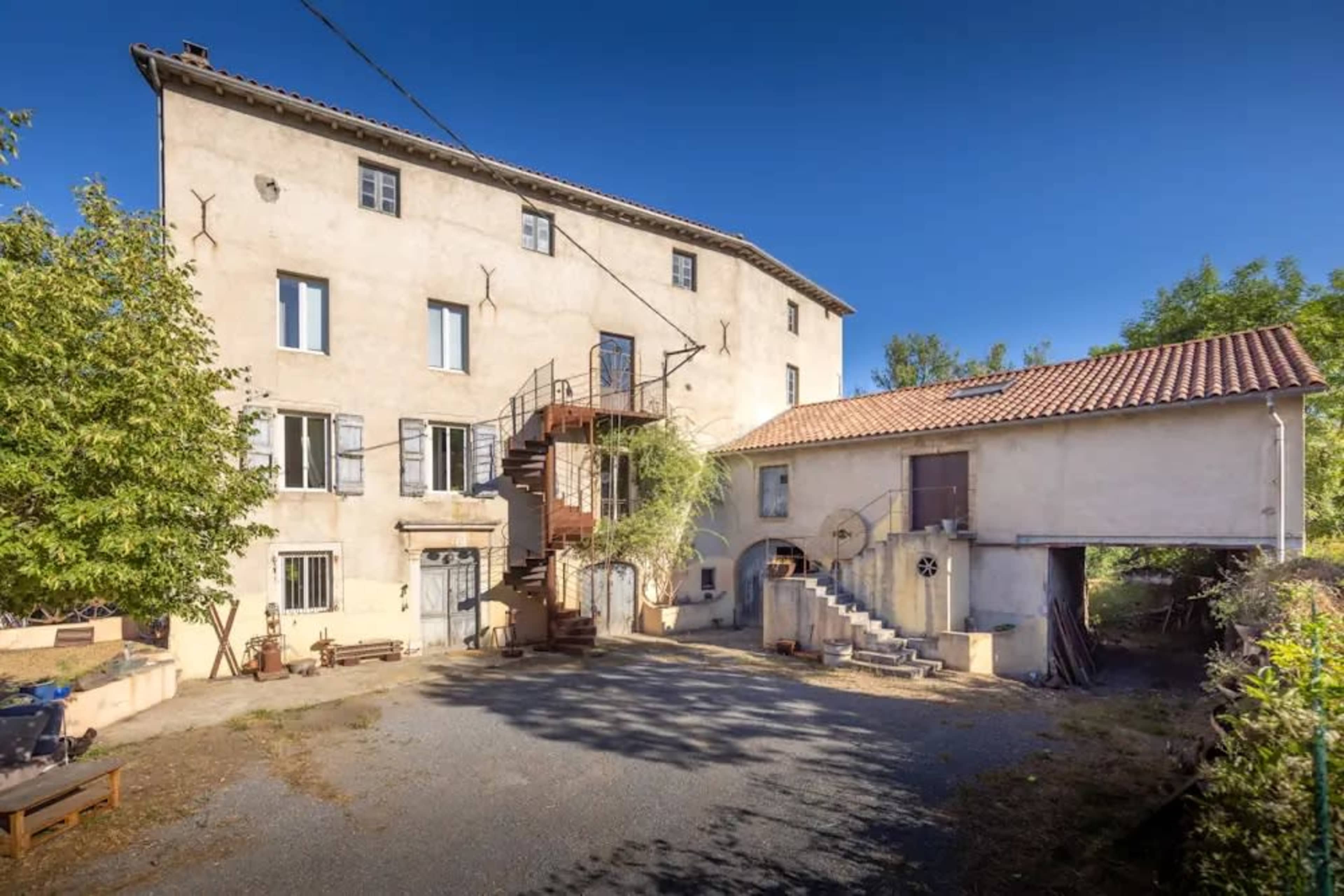 The image shows a multi-story building with a spiral staircase and a smaller adjacent structure, set in a gravel courtyard surrounded by greenery.