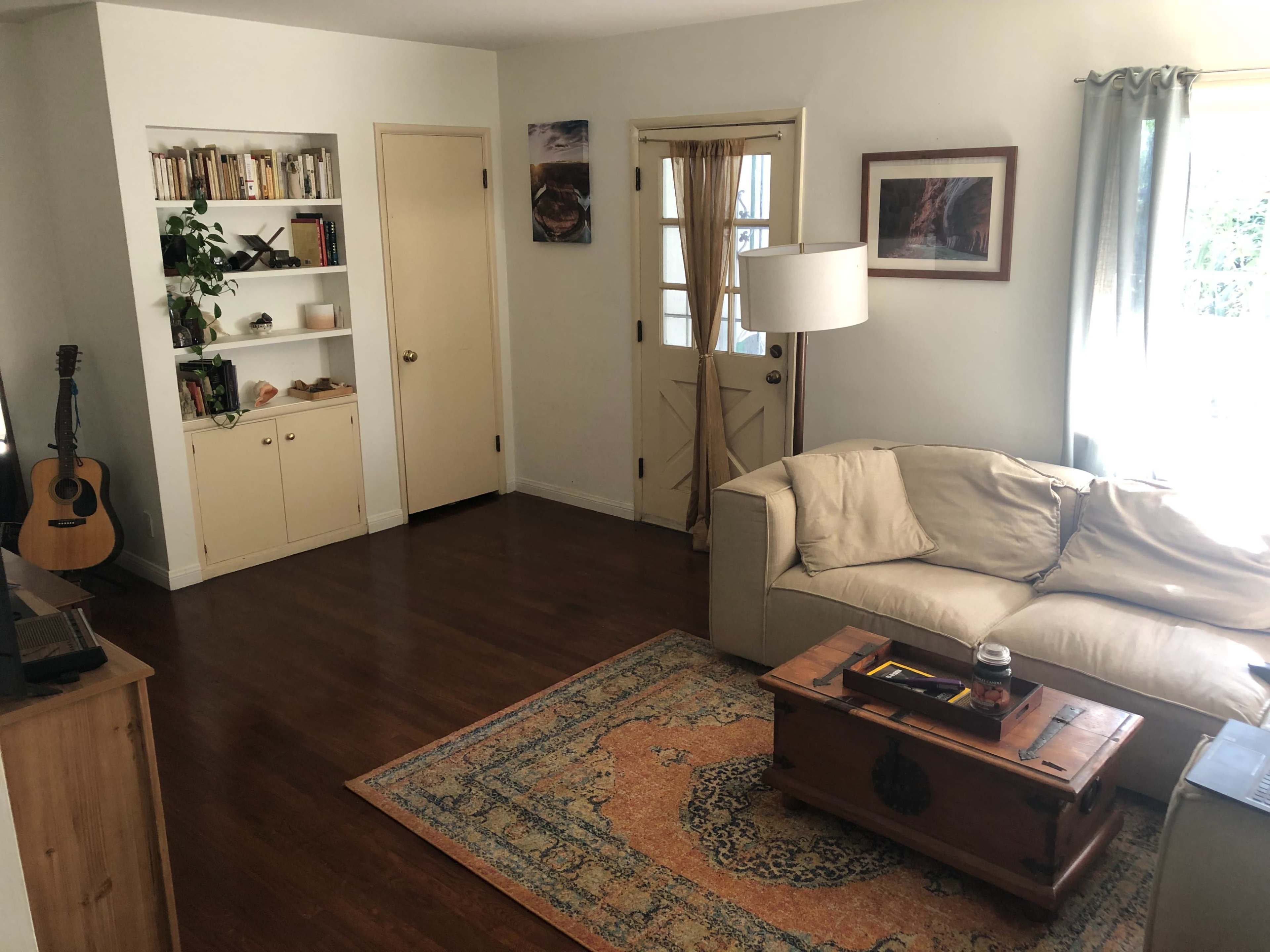 A living room with a beige couch, wooden coffee table, a bookshelf filled with books, a guitar in one corner, and a door leading outside.