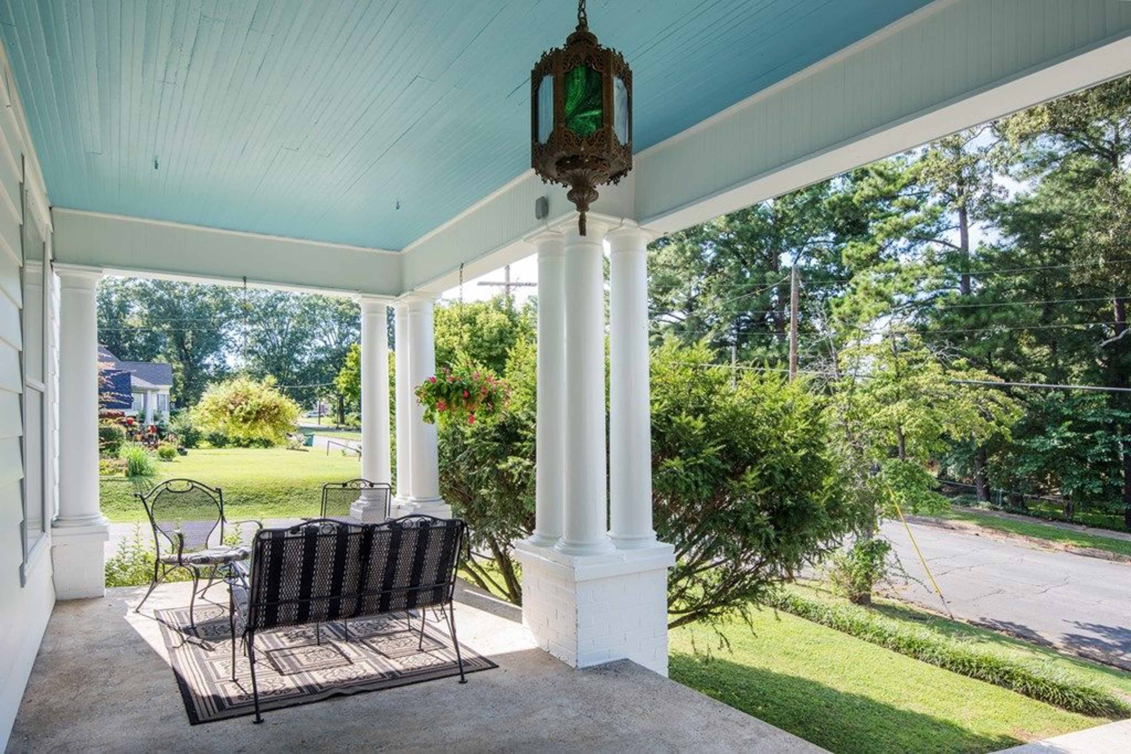 A porch with white columns, a hanging lantern, and a black metal bench overlooking a green yard and tree line.