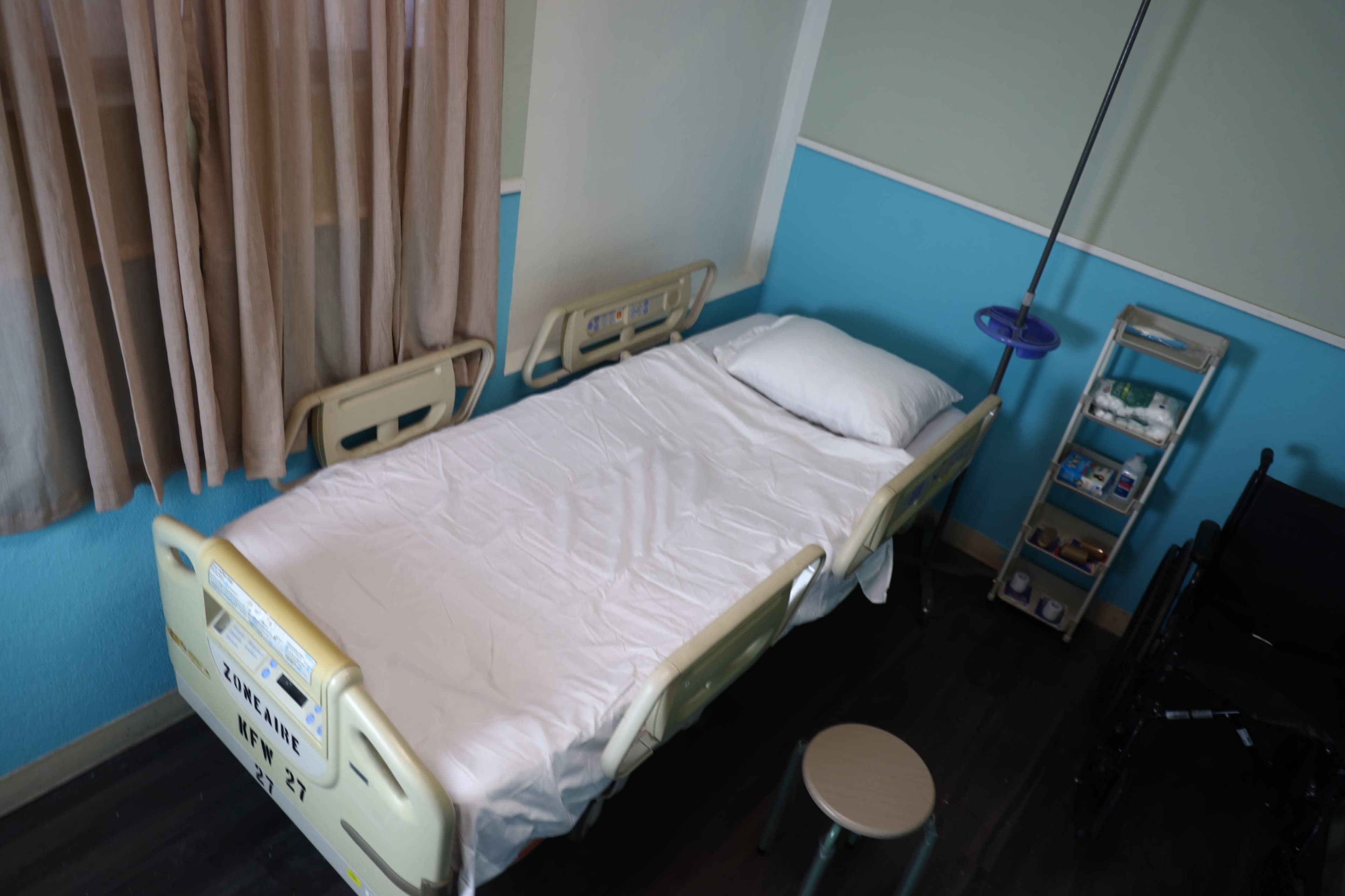 The image shows an empty hospital room with a single unoccupied bed, a stool, and a medical supply cart against a blue and white wall.