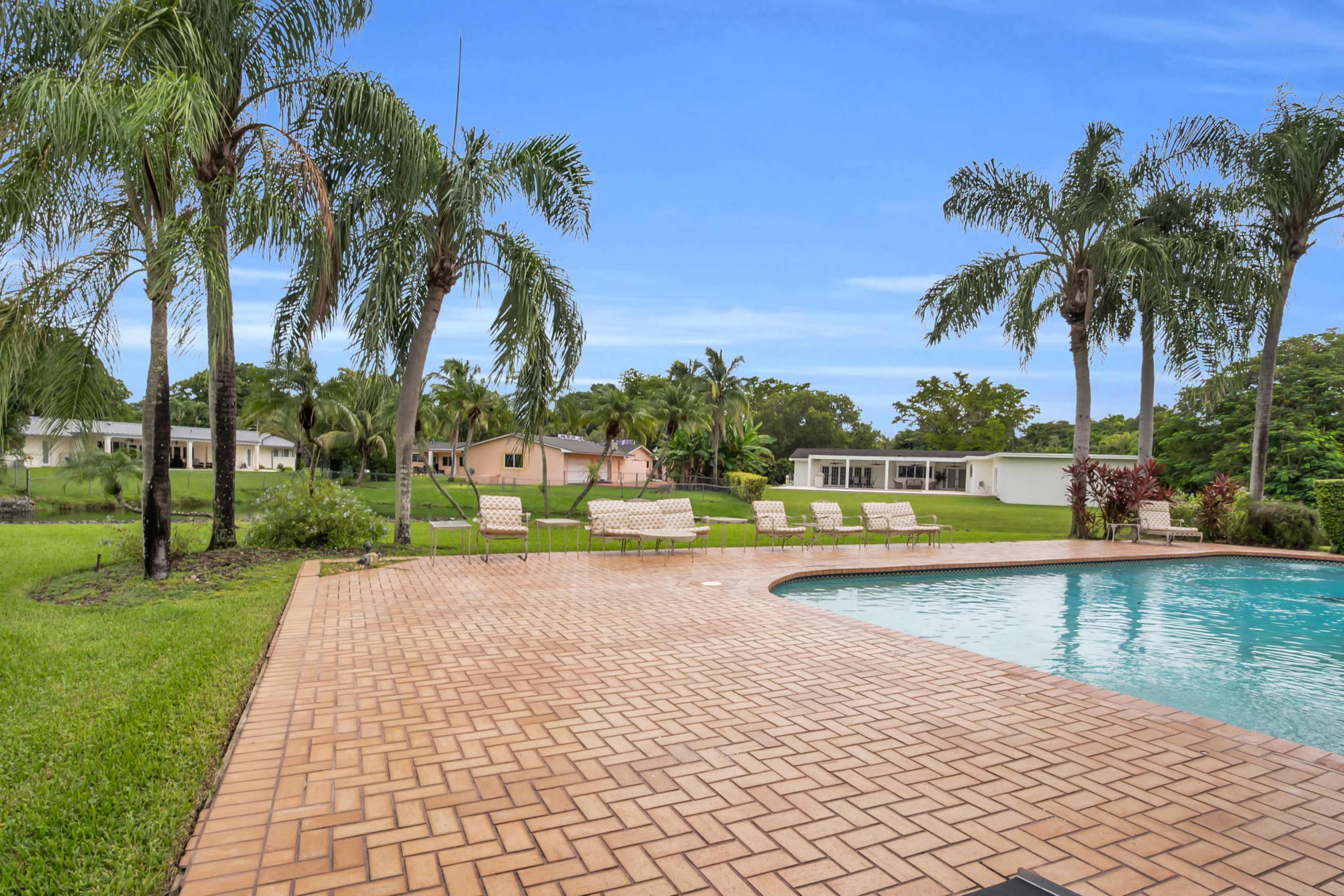 The image shows a pool surrounded by palm trees and a patio, with several lounge chairs and residential buildings in the background.