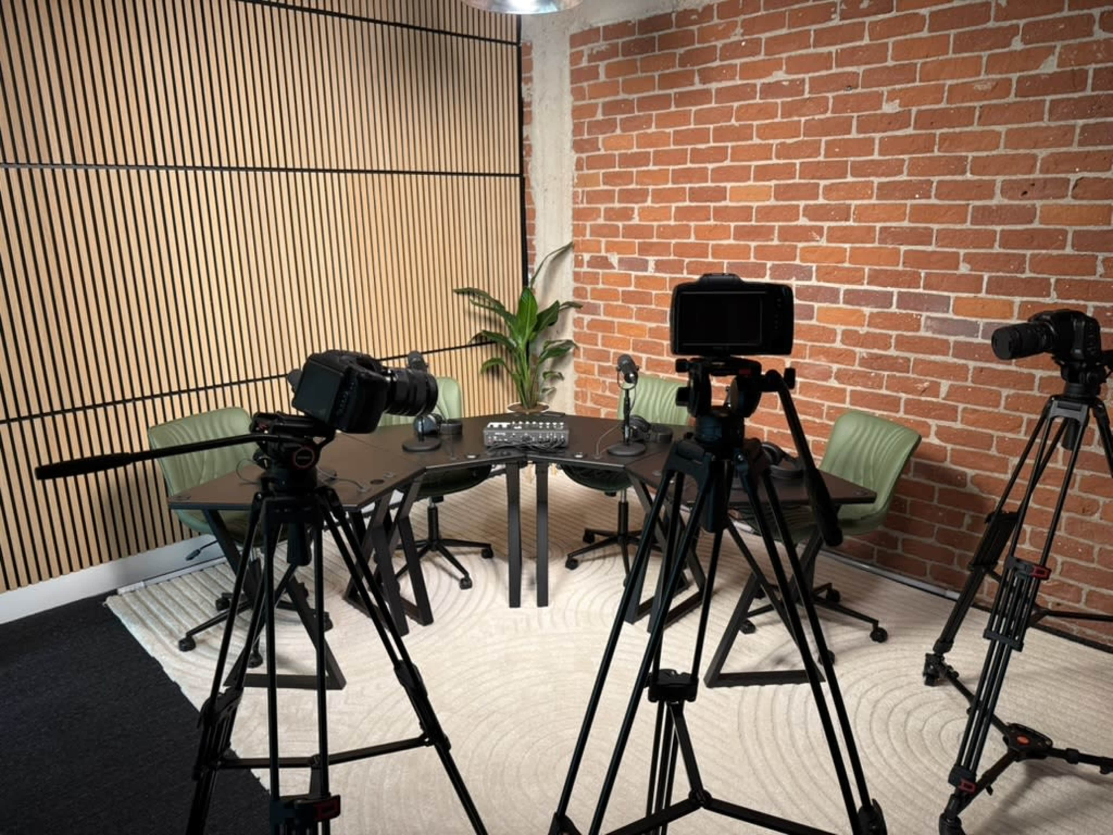 The image shows a video recording setup in a room with brick walls, featuring several cameras on tripods pointed at a round table surrounded by green chairs and a plant.