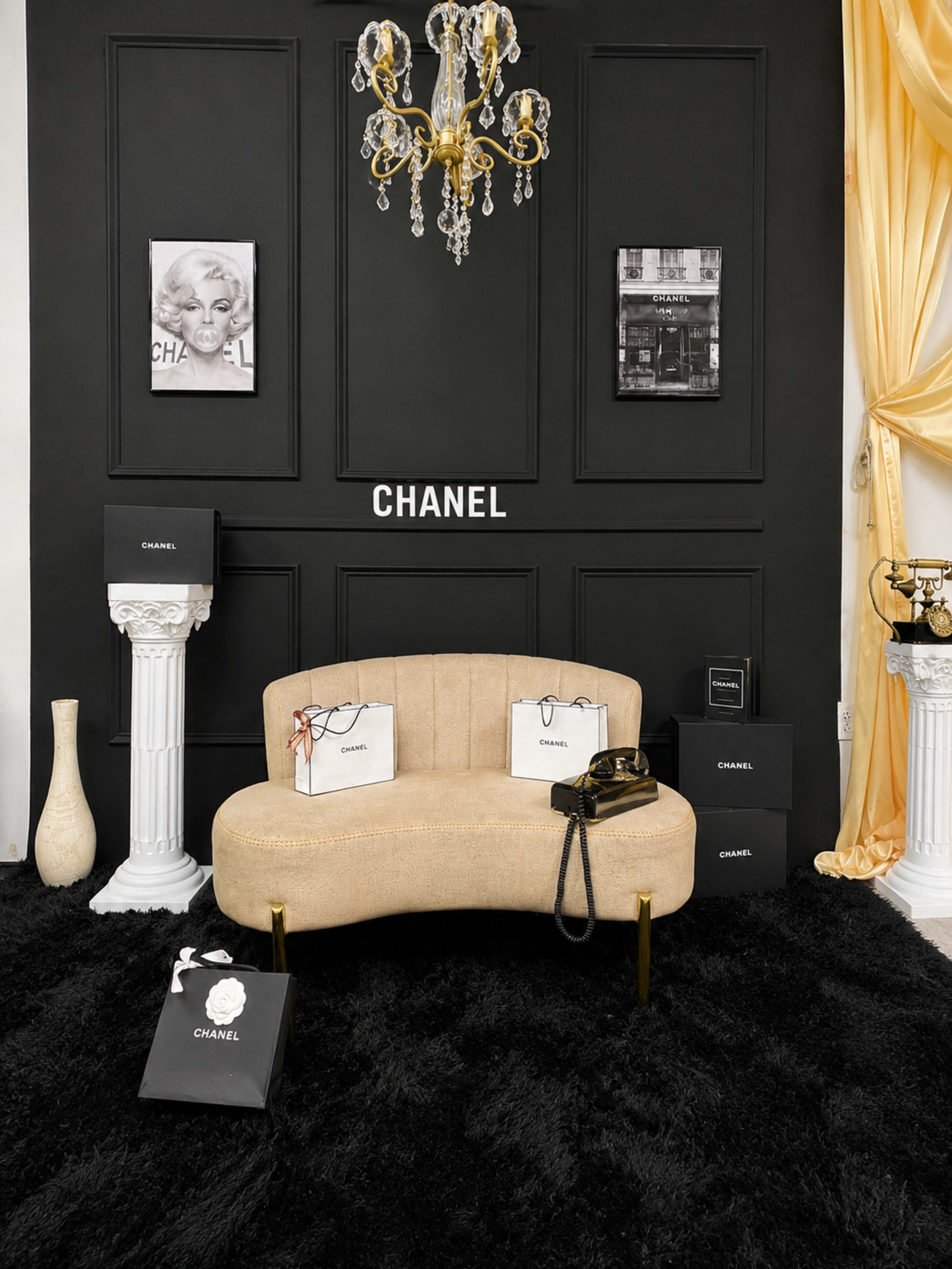 The scene features a stylish seating area with a curved beige sofa, surrounded by black Chanel shopping bags and framed art on a black wall, illuminated by a chandelier.