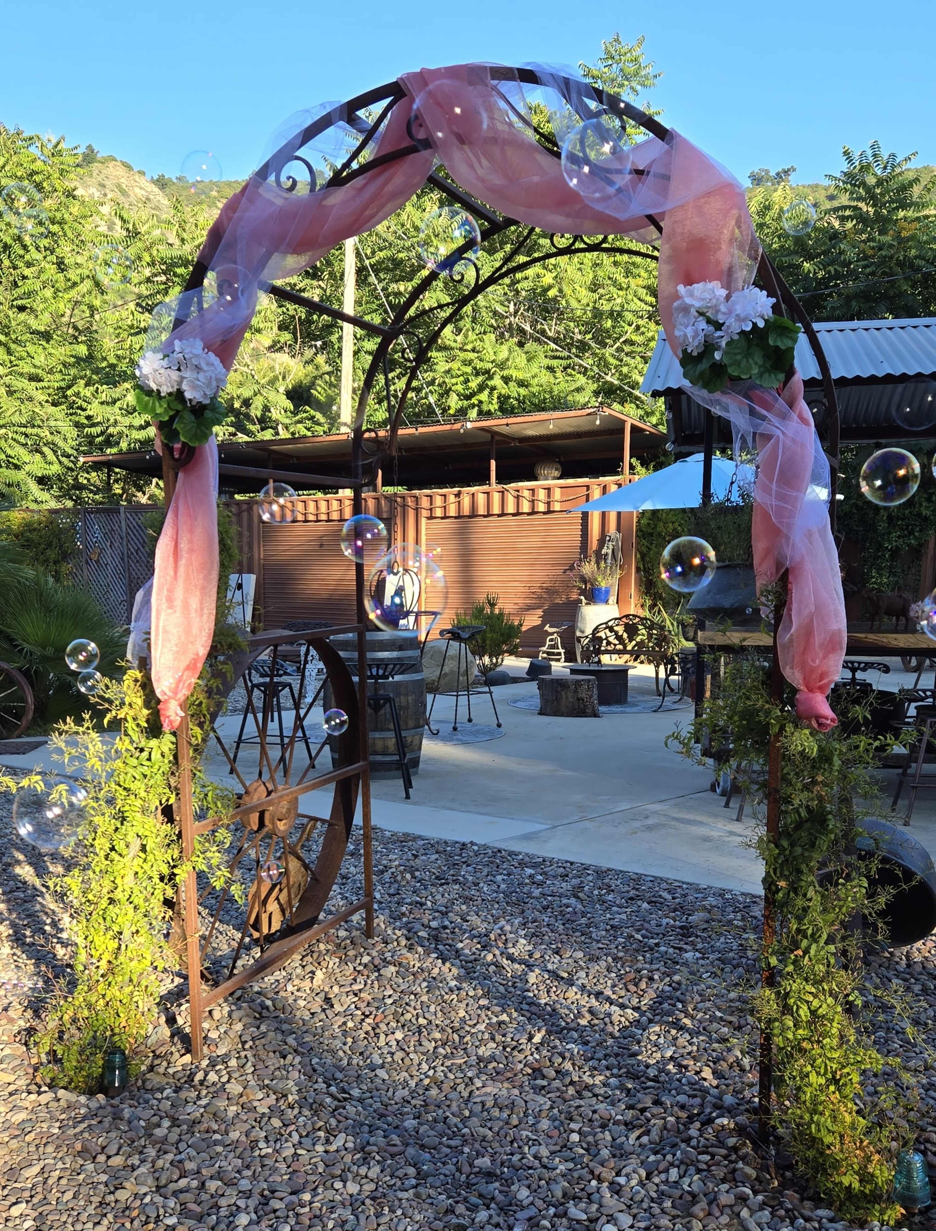 A metal arch adorned with pink fabric and flowers, surrounded by bubbles, stands in a gravel area with seating and greenery in the background.