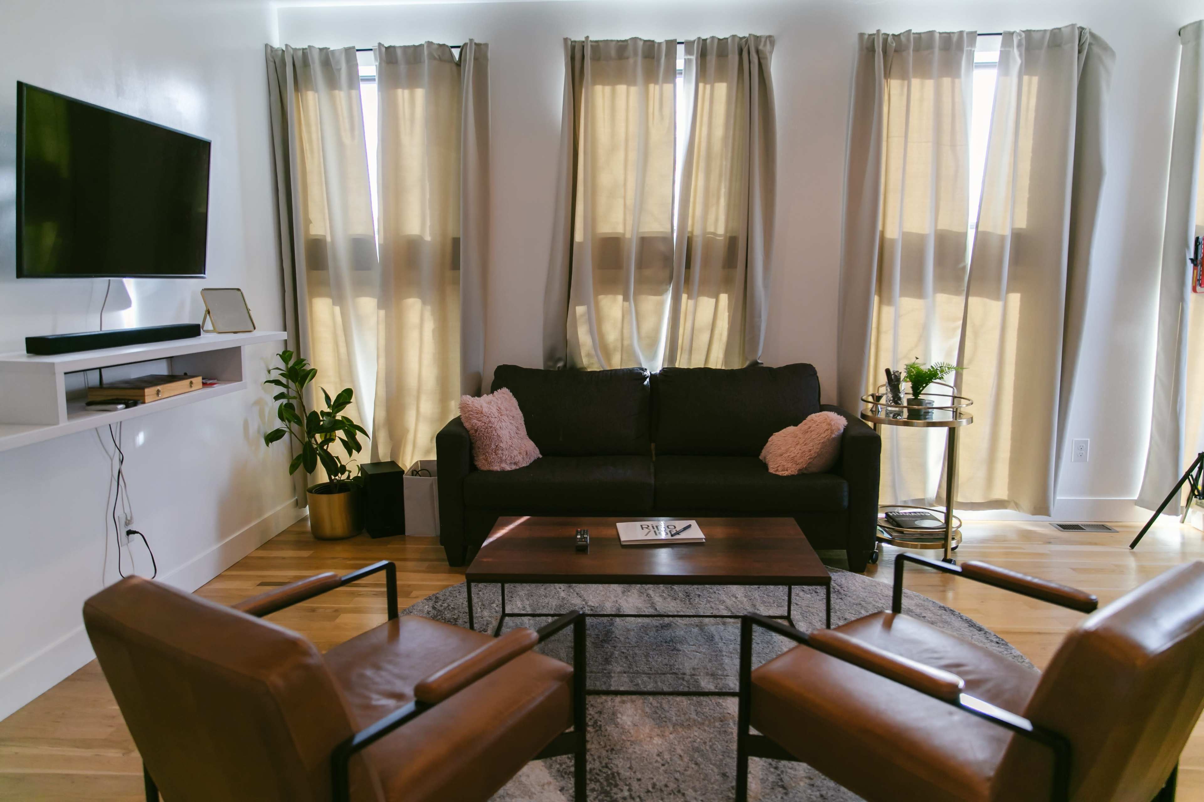 A modern living room features a dark sofa with cushions, two leather chairs, a coffee table, and large windows with light drapes.