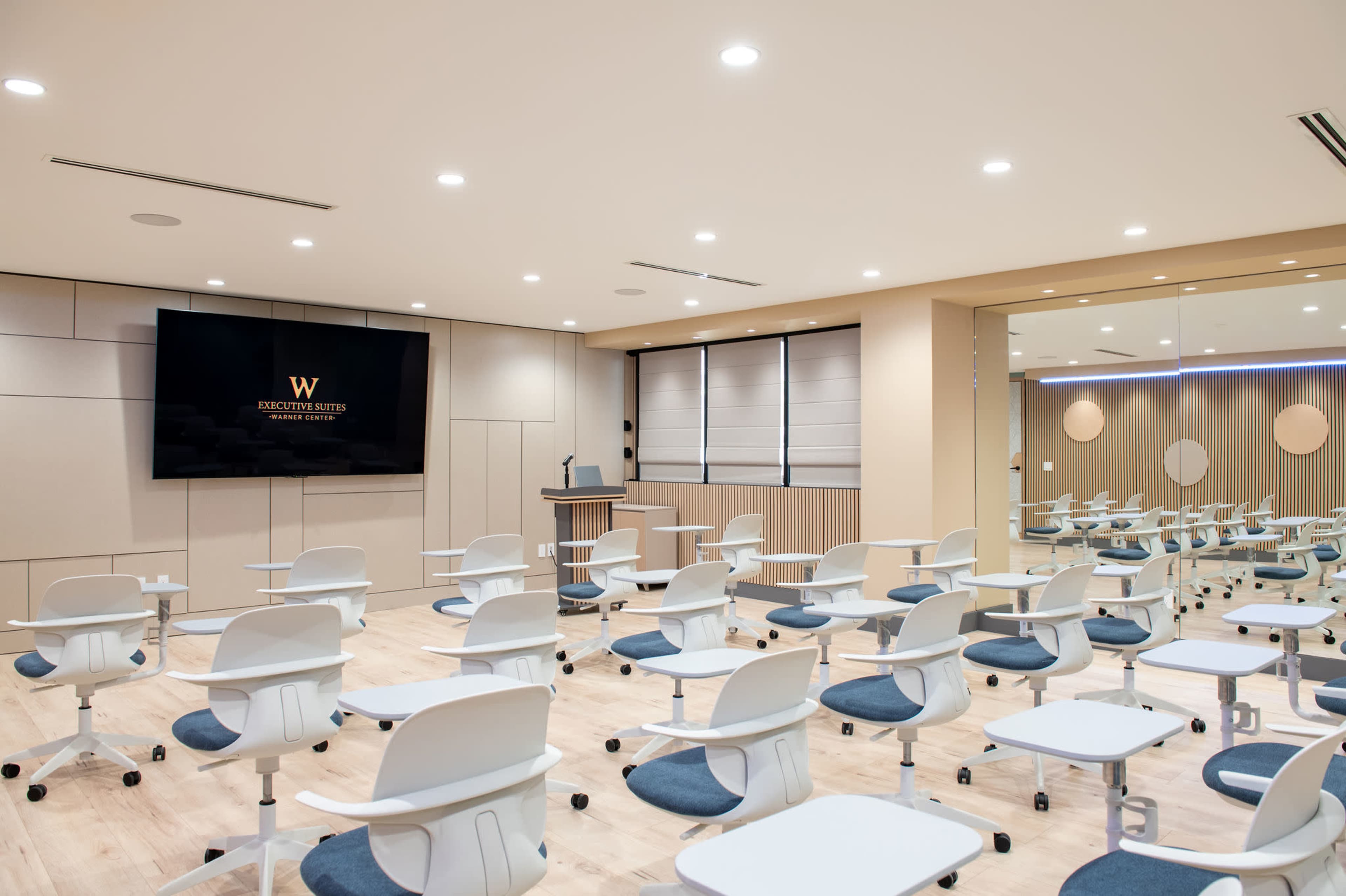 The image shows a modern classroom setup with rows of movable chairs and tables facing a large screen displaying a logo on the front wall.