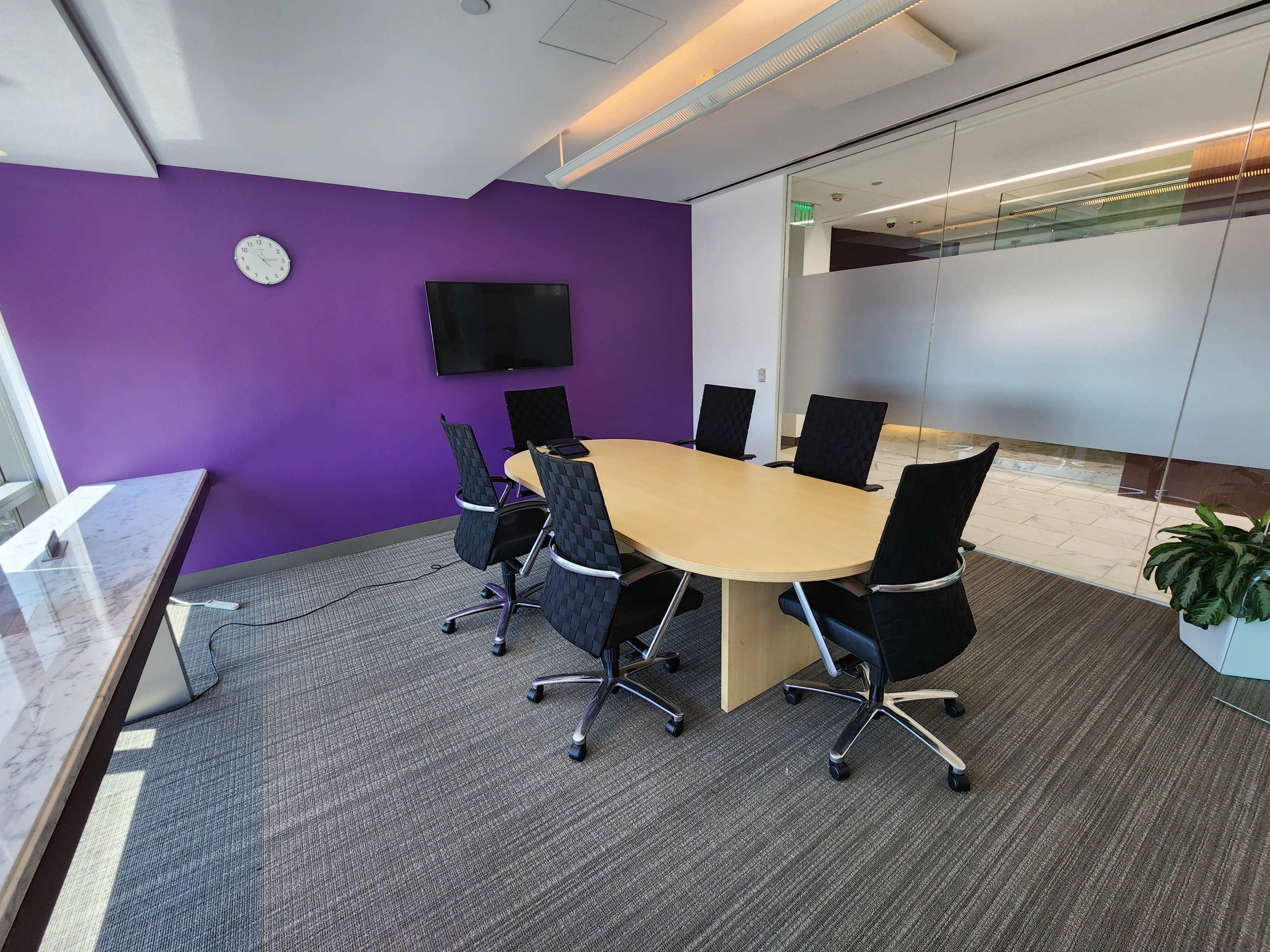 A conference room features a round wooden table surrounded by six black chairs, with a purple wall and a wall-mounted TV visible in the background.