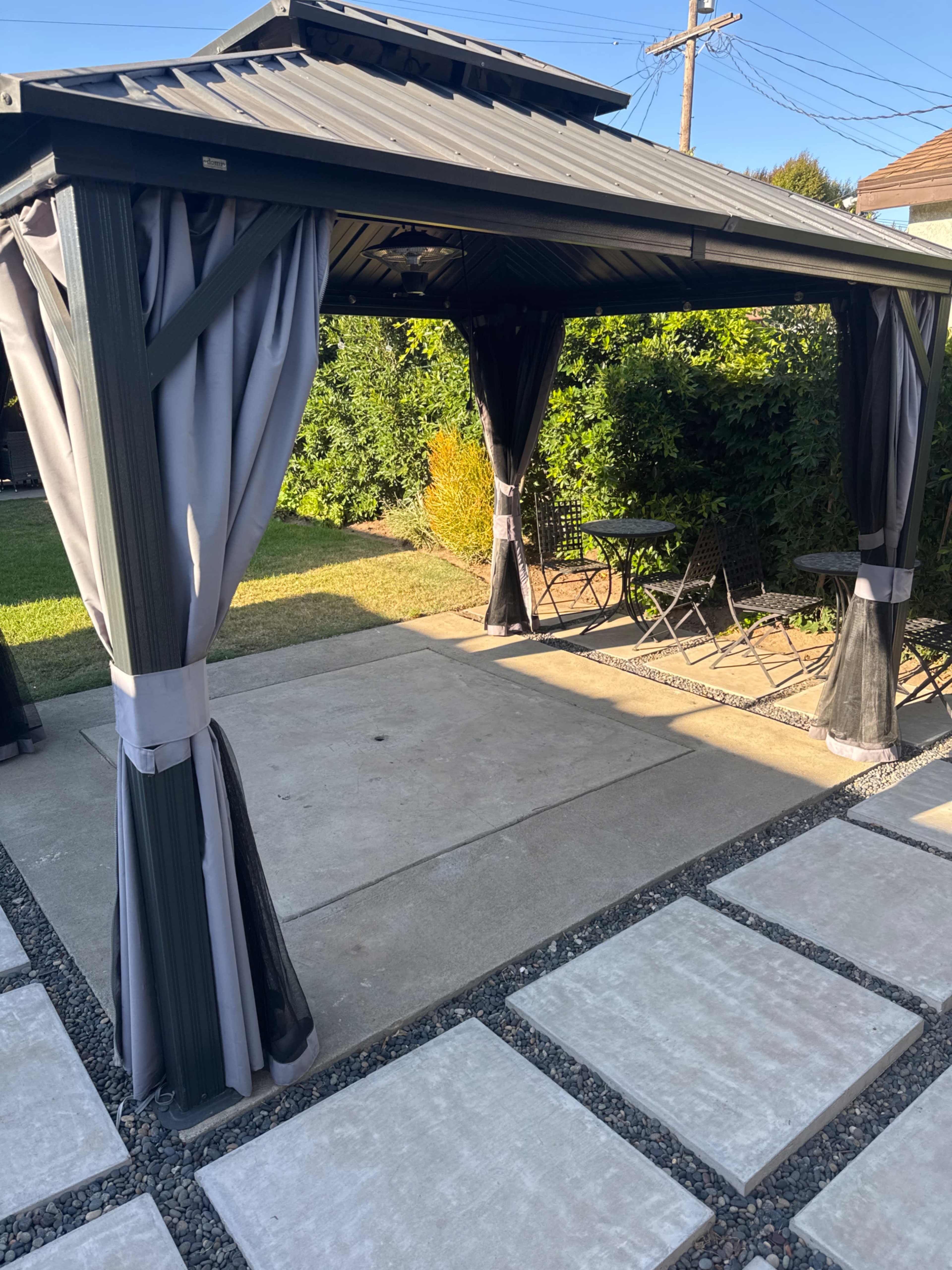 A metal gazebo with fabric curtains overlooks a stone pathway leading to a small seating area with black chairs and a table.