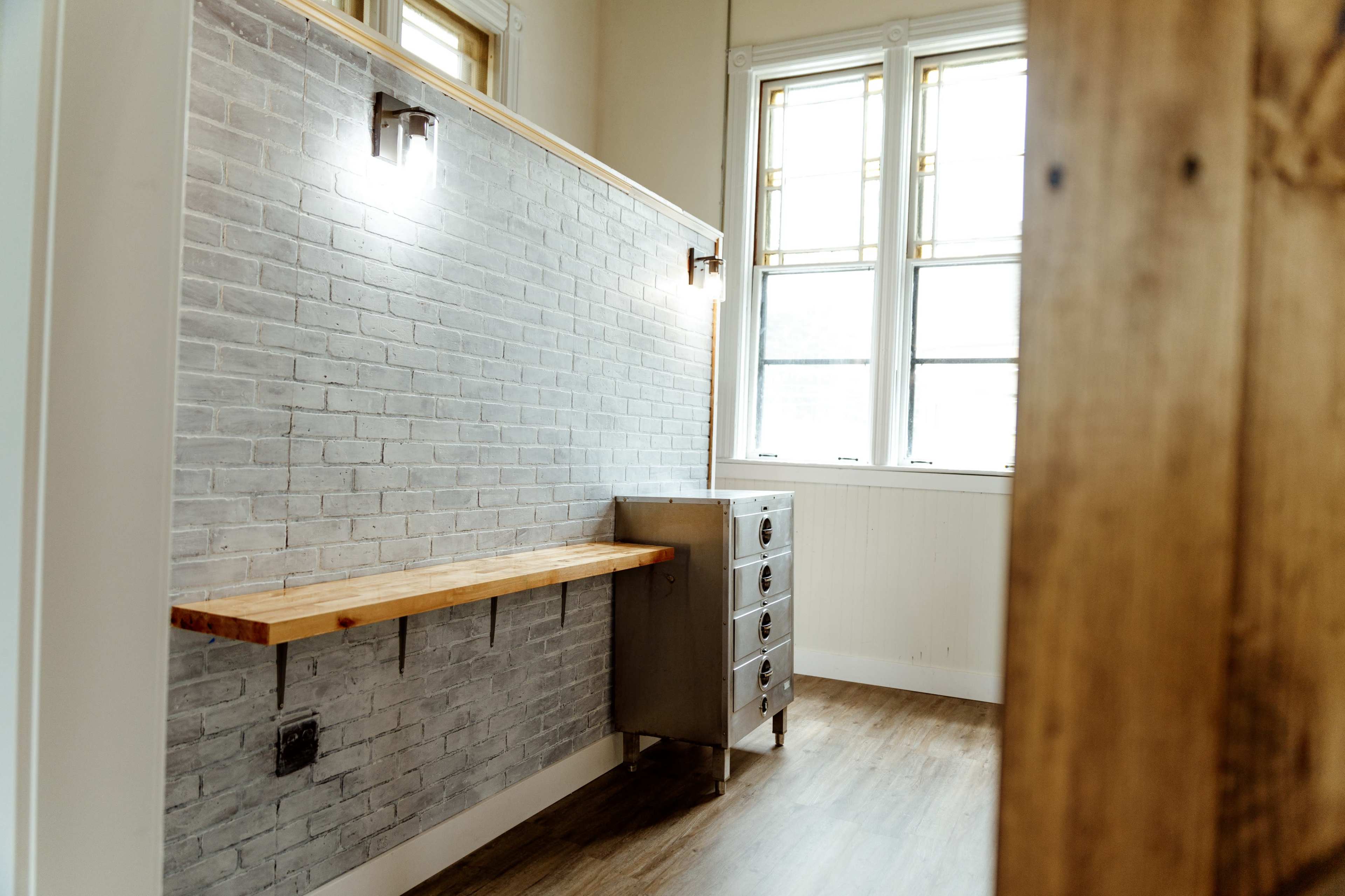 The image shows a room with a wooden counter along a gray brick wall, alongside a metal drawer unit, illuminated by wall sconces and natural light from large windows.