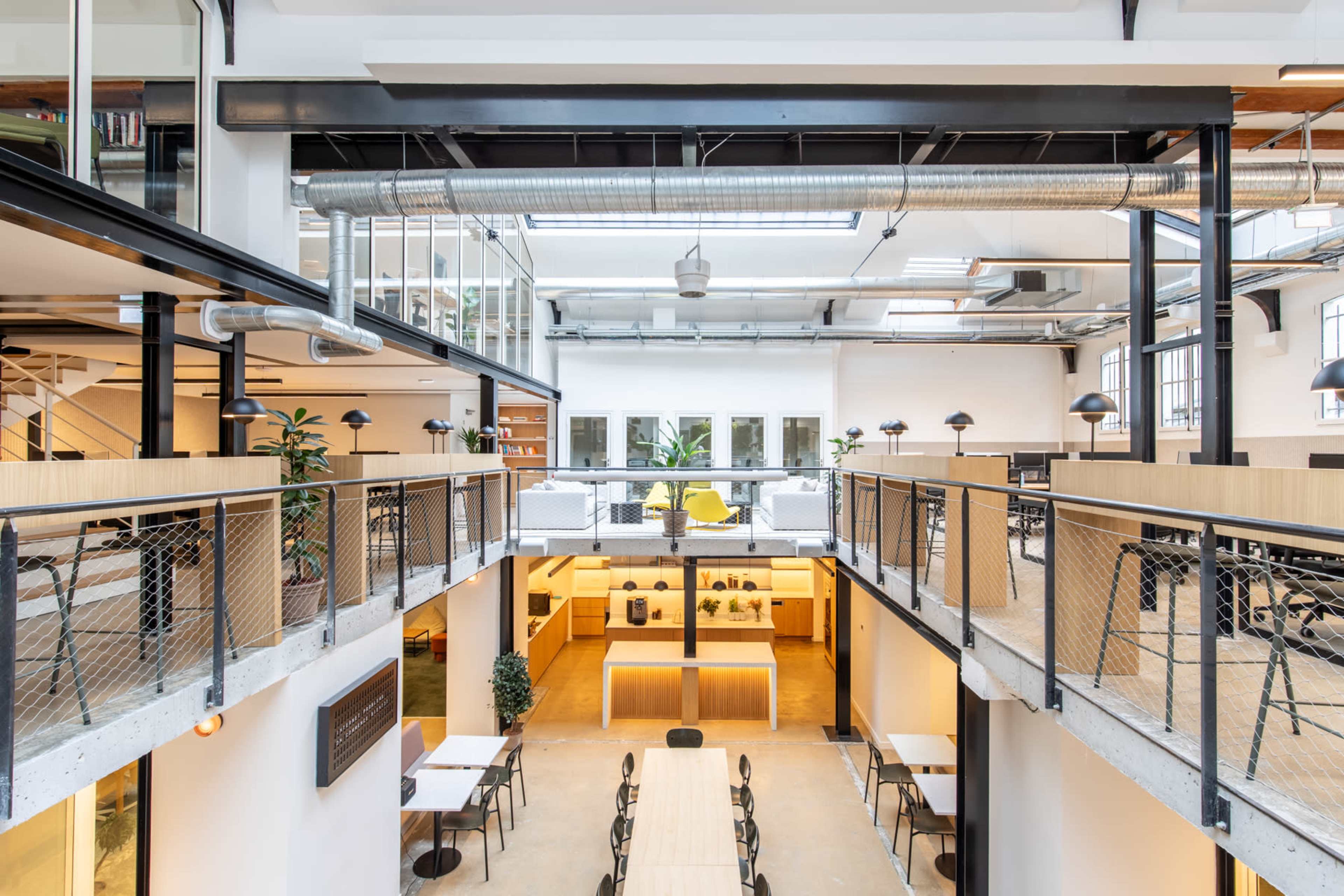 An industrial-style open-plan office with multiple levels, featuring wooden furniture, greenery, and large windows allowing natural light.