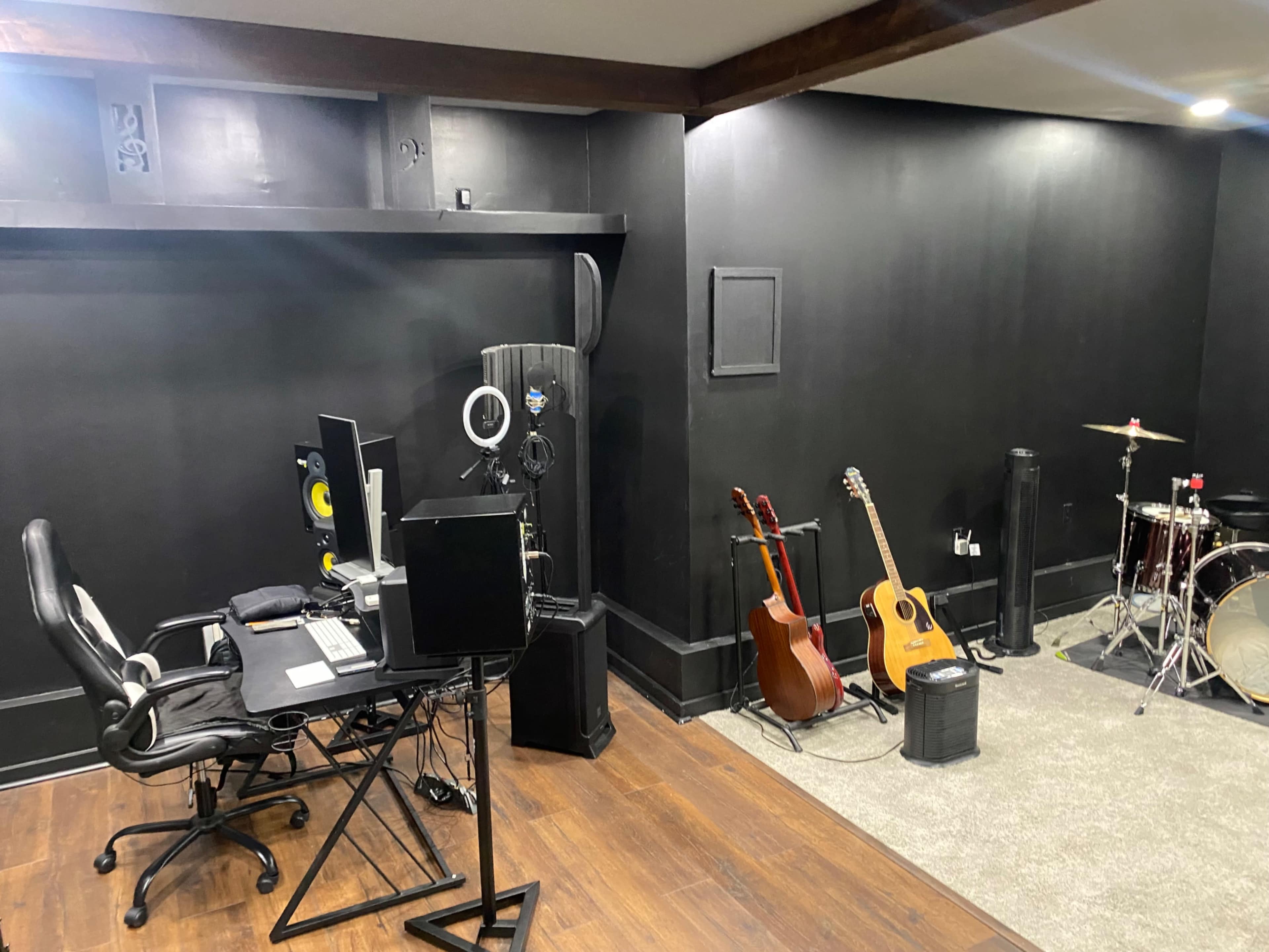 The image shows a music studio with a desk featuring computer equipment, two guitars propped against the wall, and a drum set in the corner.