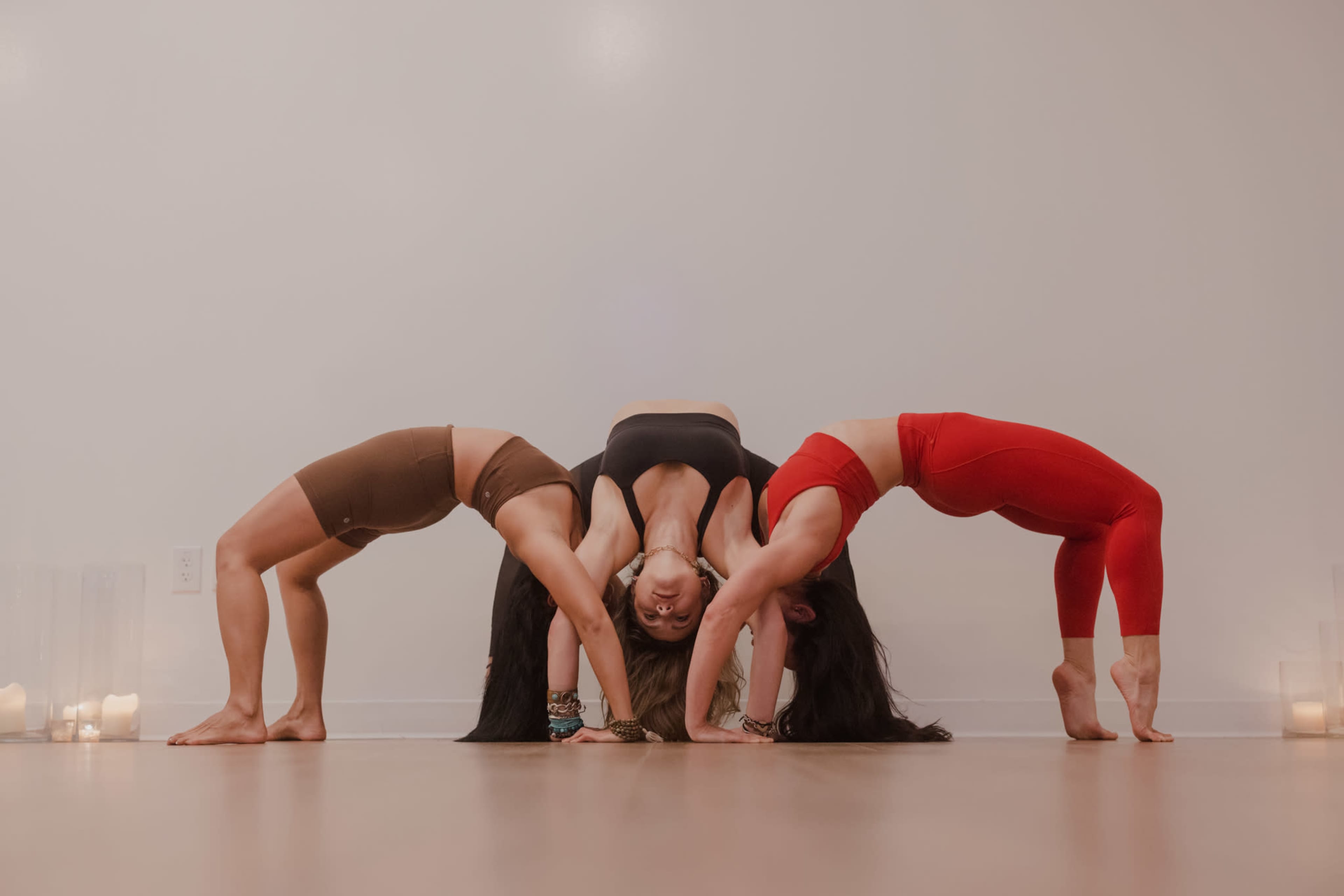 Three women perform a yoga pose in a studio, forming an arch with their bodies, while candles are lit in the background.