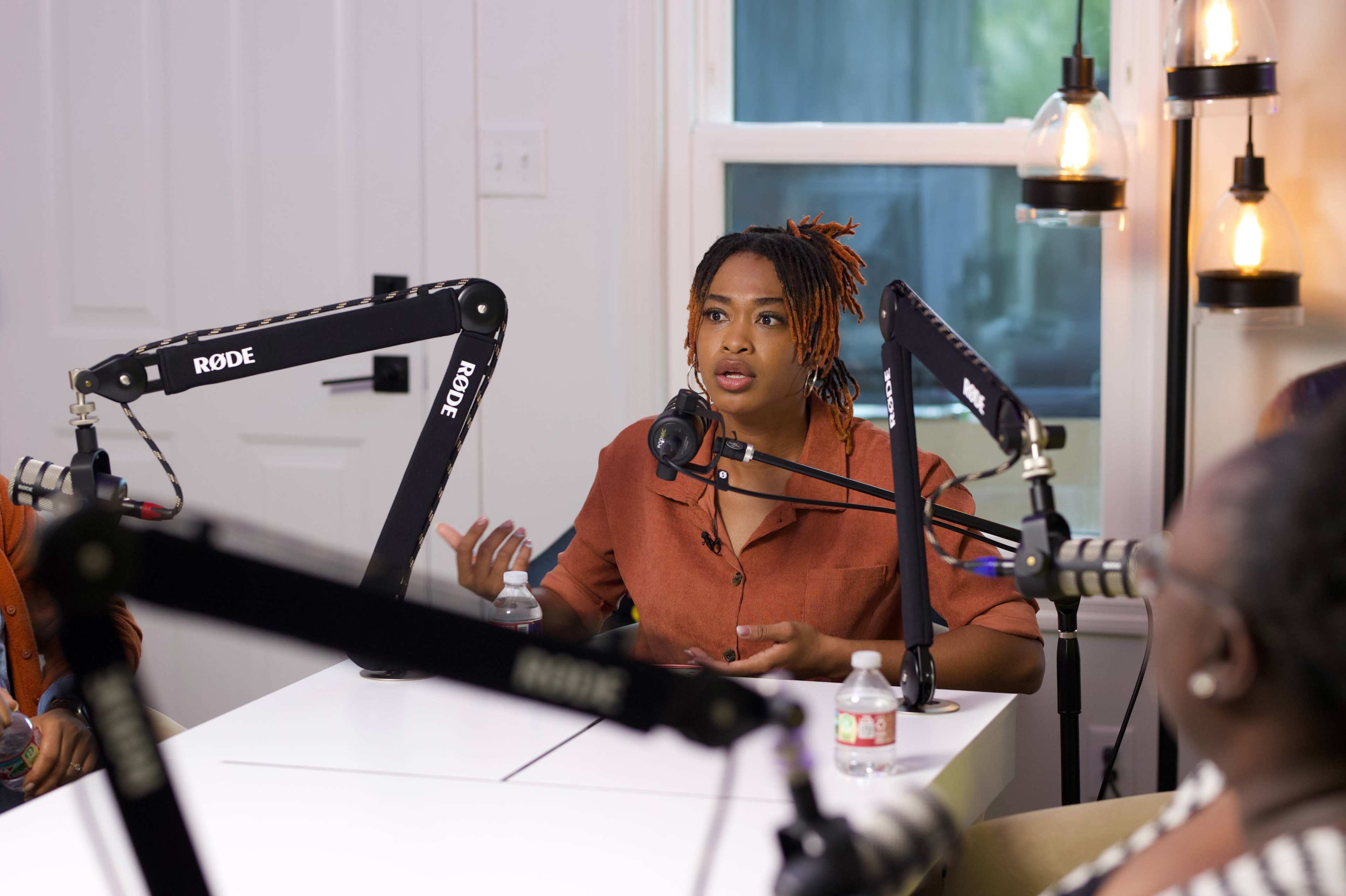A woman with braided hair talks animatedly into a microphone at a podcast recording setup with bright lighting and a modern aesthetic.