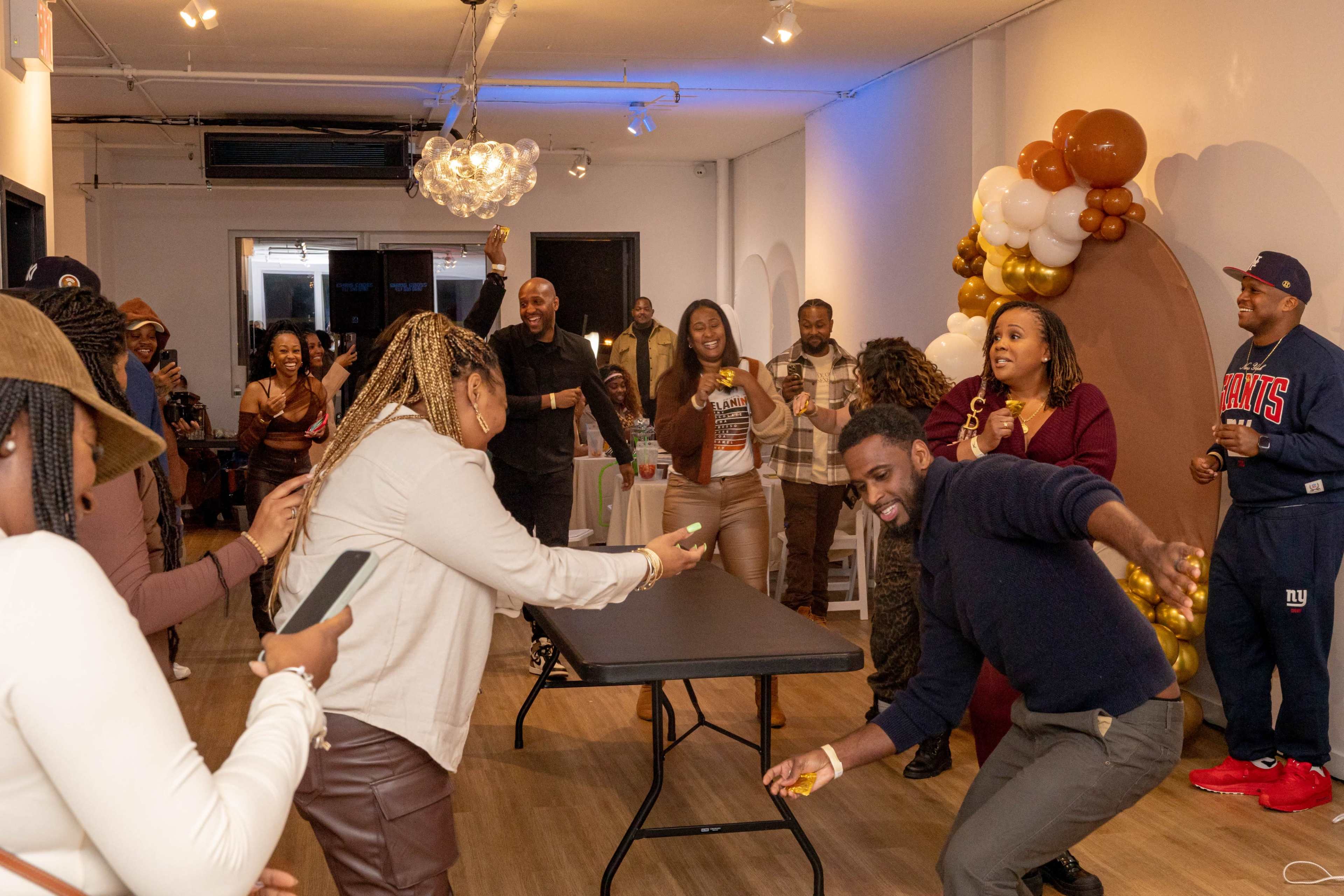 A group of people is gathered in a brightly lit room, participating in a game around a table decorated for a celebration.