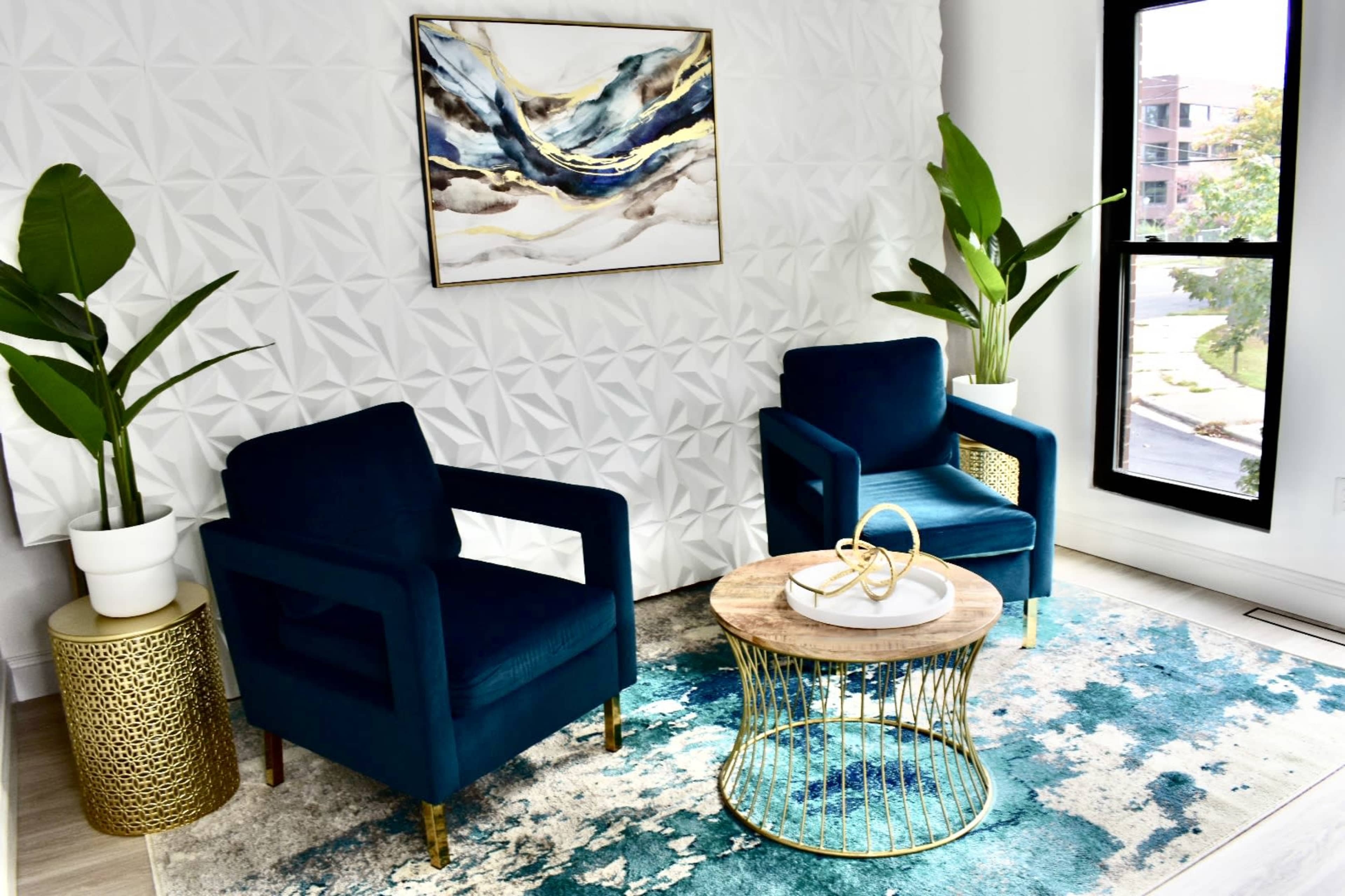 The image shows a modern living space featuring two blue chairs, a round wooden coffee table, and large plants, all arranged on a patterned rug in front of a textured white wall.
