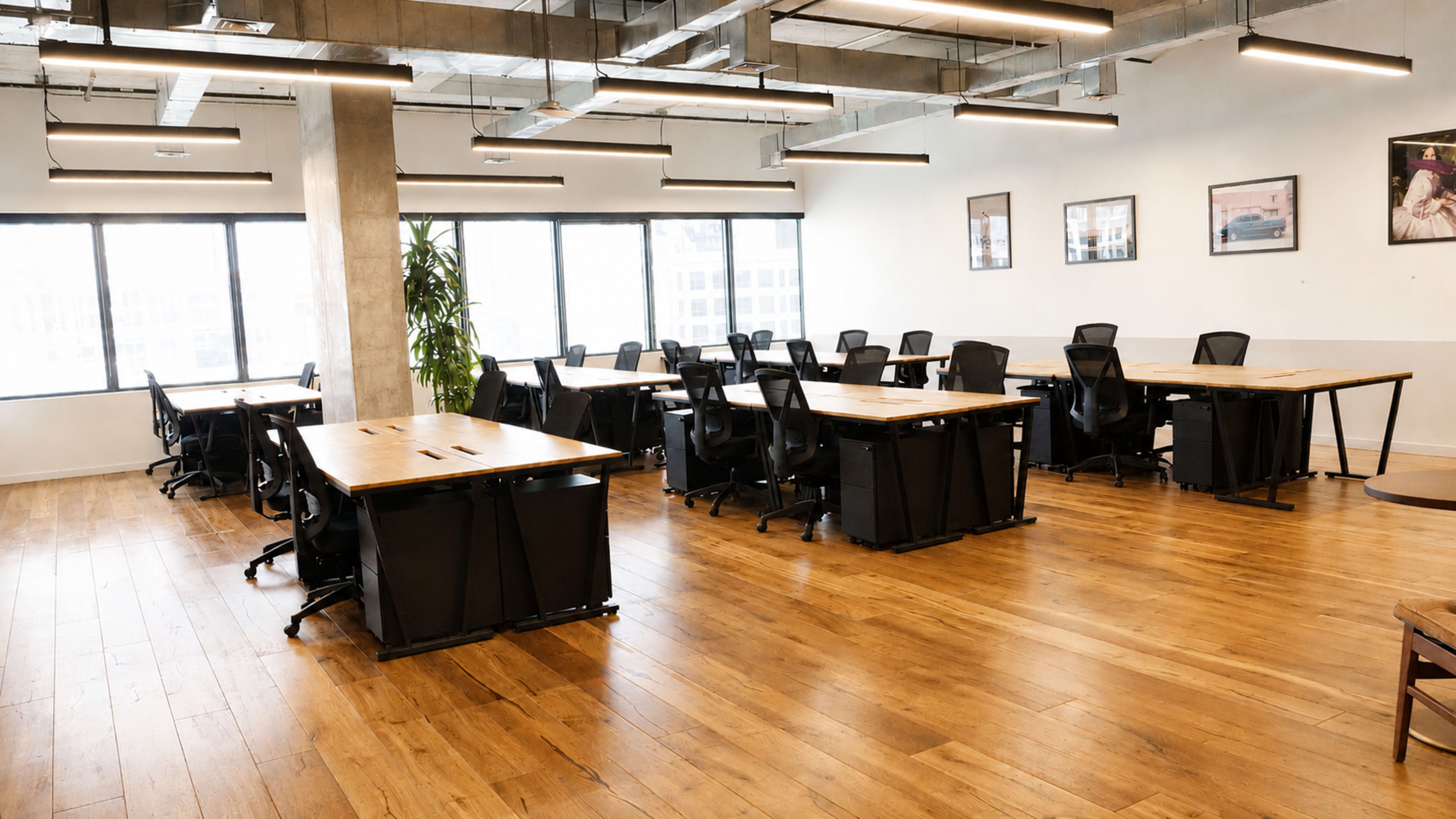 The image shows a modern office space with multiple wooden desks and ergonomic chairs arranged in a well-lit room featuring large windows and a wooden floor.