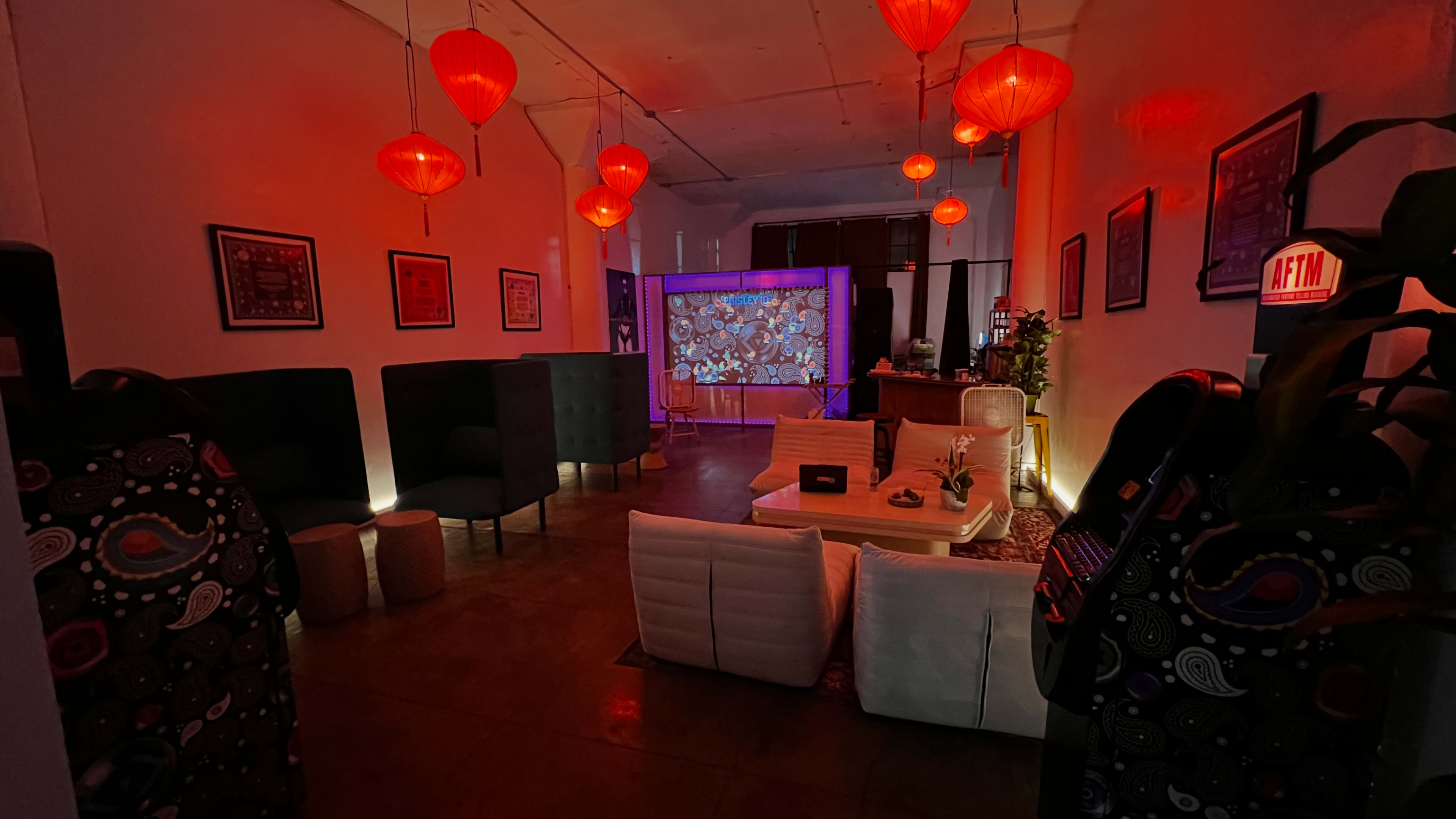 The image shows a room with red lanterns hanging from the ceiling, a large projection screen displaying colorful visuals, and seating areas arranged around a central coffee table.