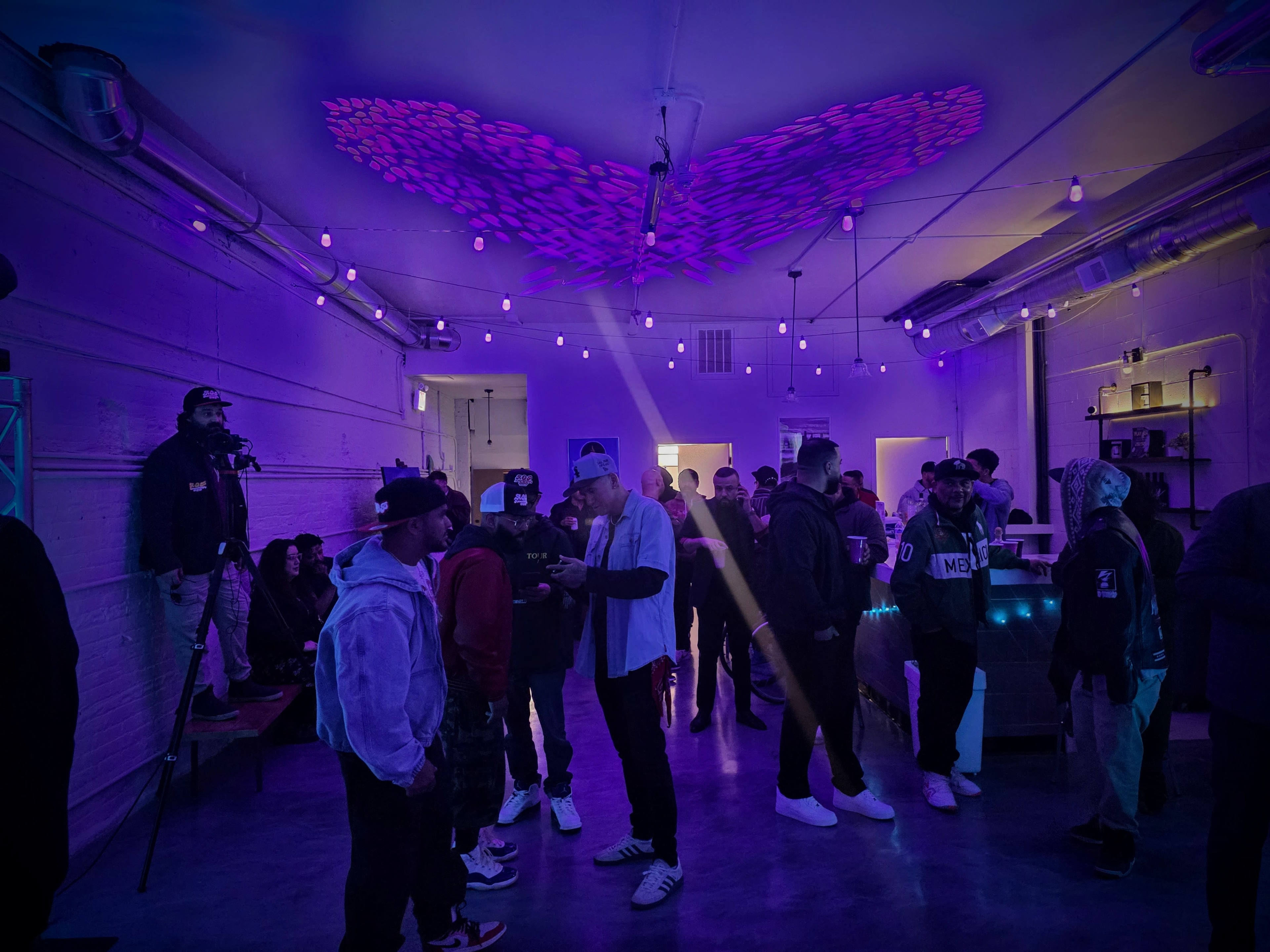A group of people socialize in a dimly lit purple-themed space with string lights overhead and a decorative light pattern on the ceiling.