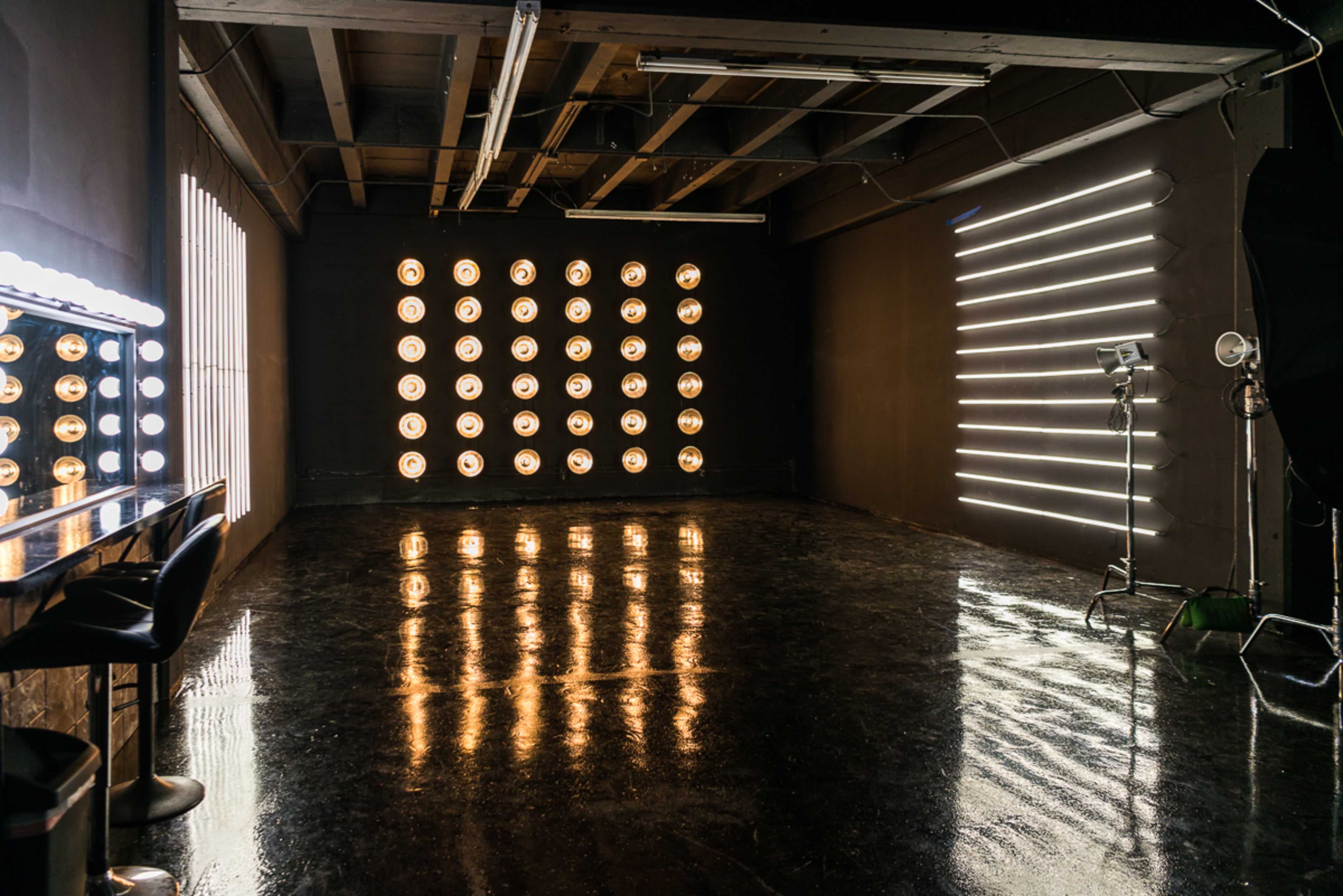 The image shows a photography studio with illuminated circular lights and vertical light strips on two walls, along with a mirror and stools on one side.