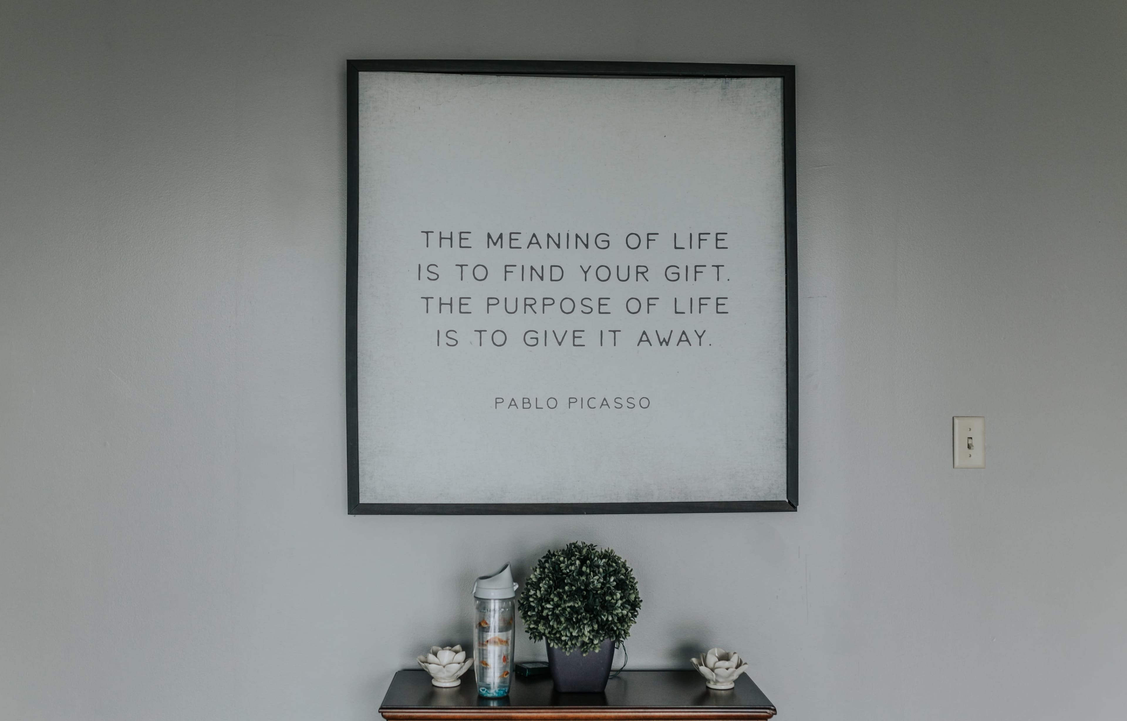A framed quote by Pablo Picasso about the meaning and purpose of life is displayed on a wall above a small table with a plant and decorative items.