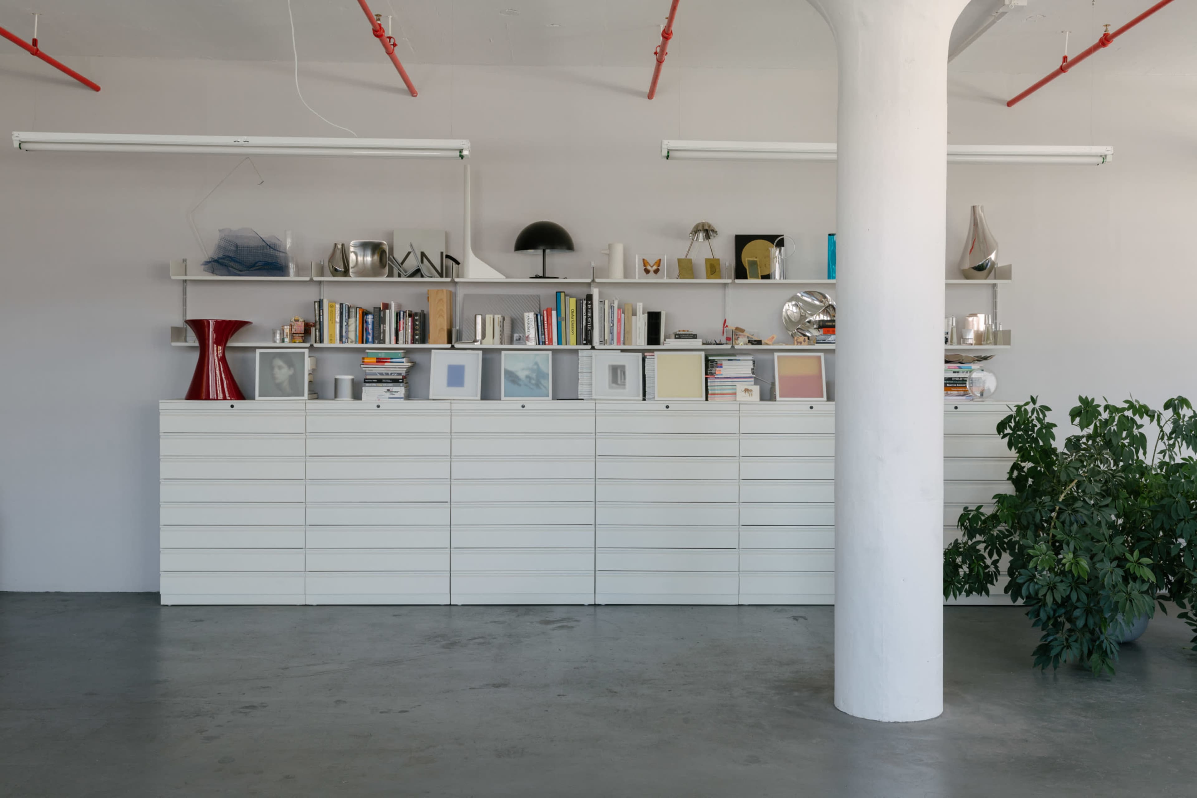 The image shows a modern, minimalist room featuring a long white storage unit against a light gray wall, adorned with books and decorative objects on a shelf above it, with a green plant in the foreground and a column in the center.