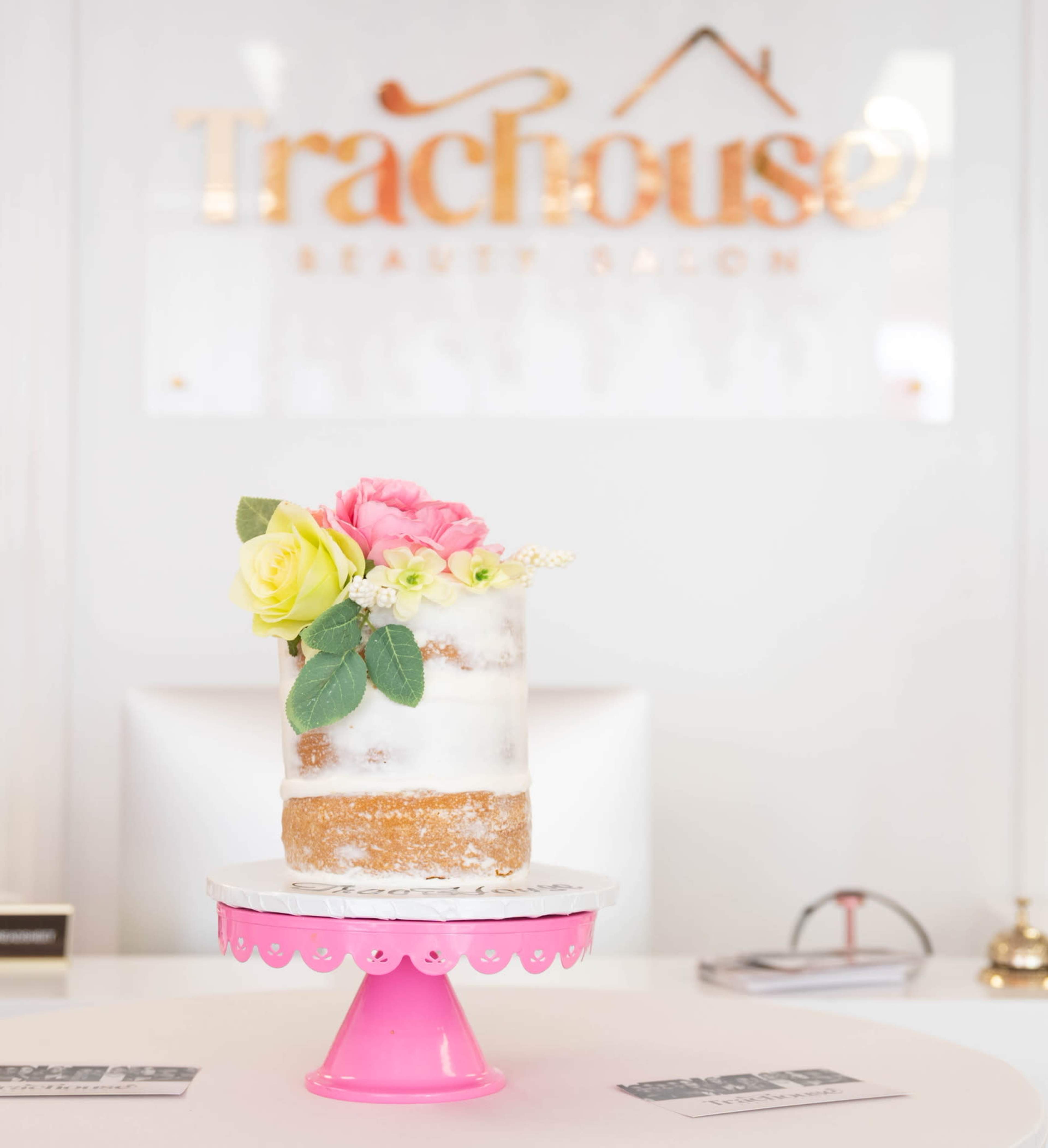 A decorated cake topped with flowers is displayed on a pink pedestal in a beauty salon setting.