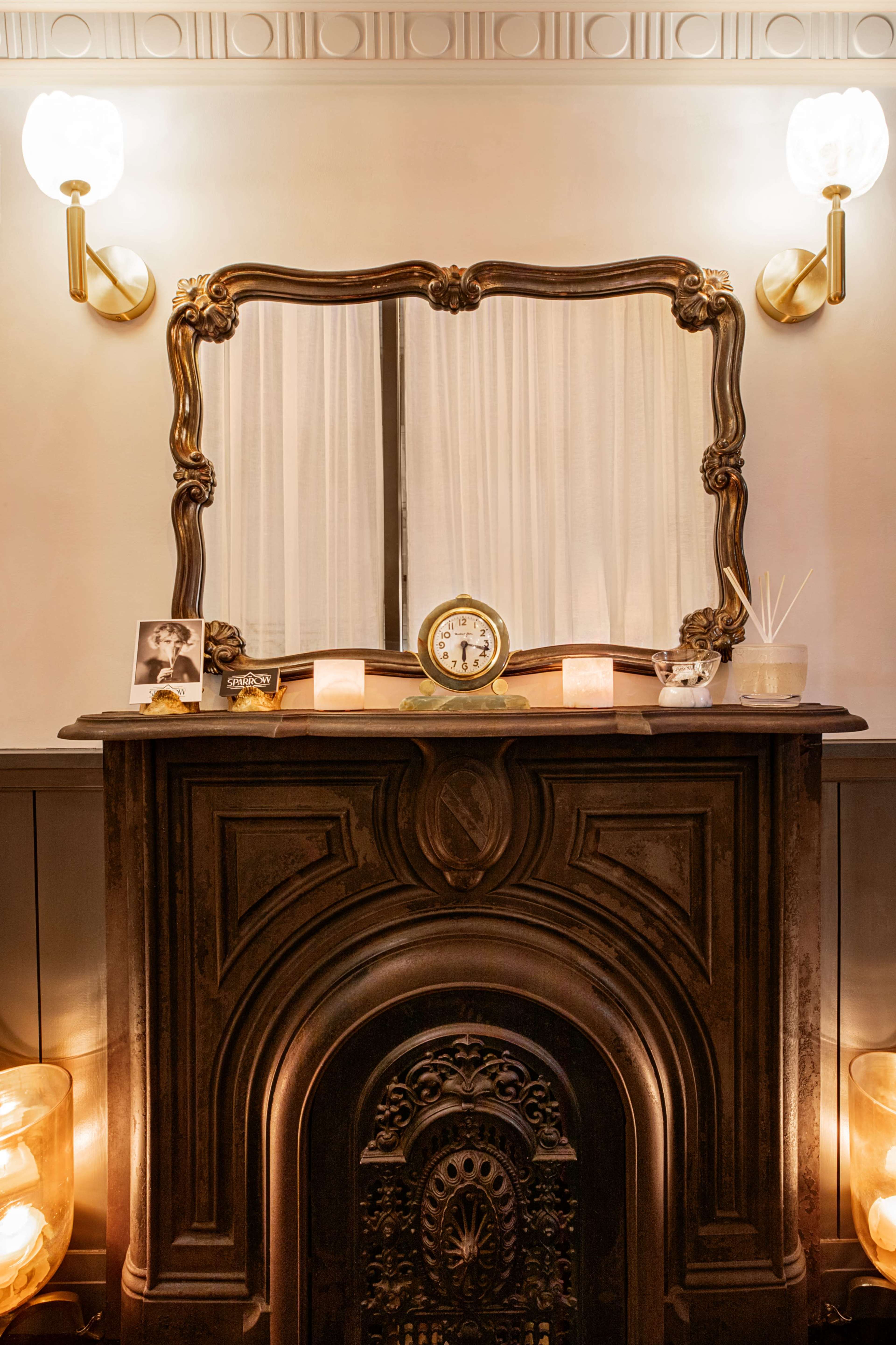 A vintage fireplace is decorated with a large ornate mirror, a clock, and a few decorative items, illuminated by wall sconces and warm light sources.