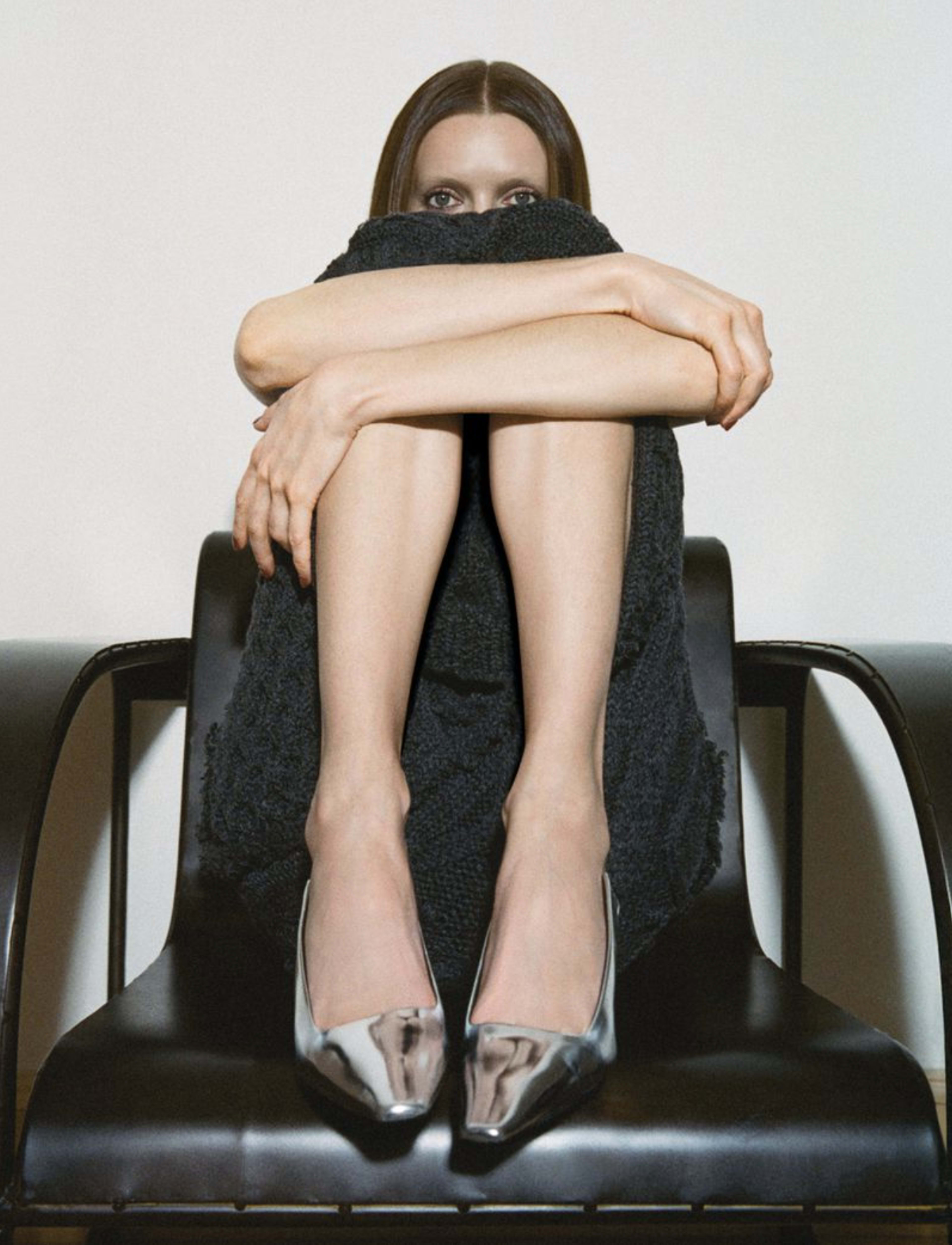 A person with long hair sits on a chair, hugging their knees, and wearing shiny silver high-heeled shoes.