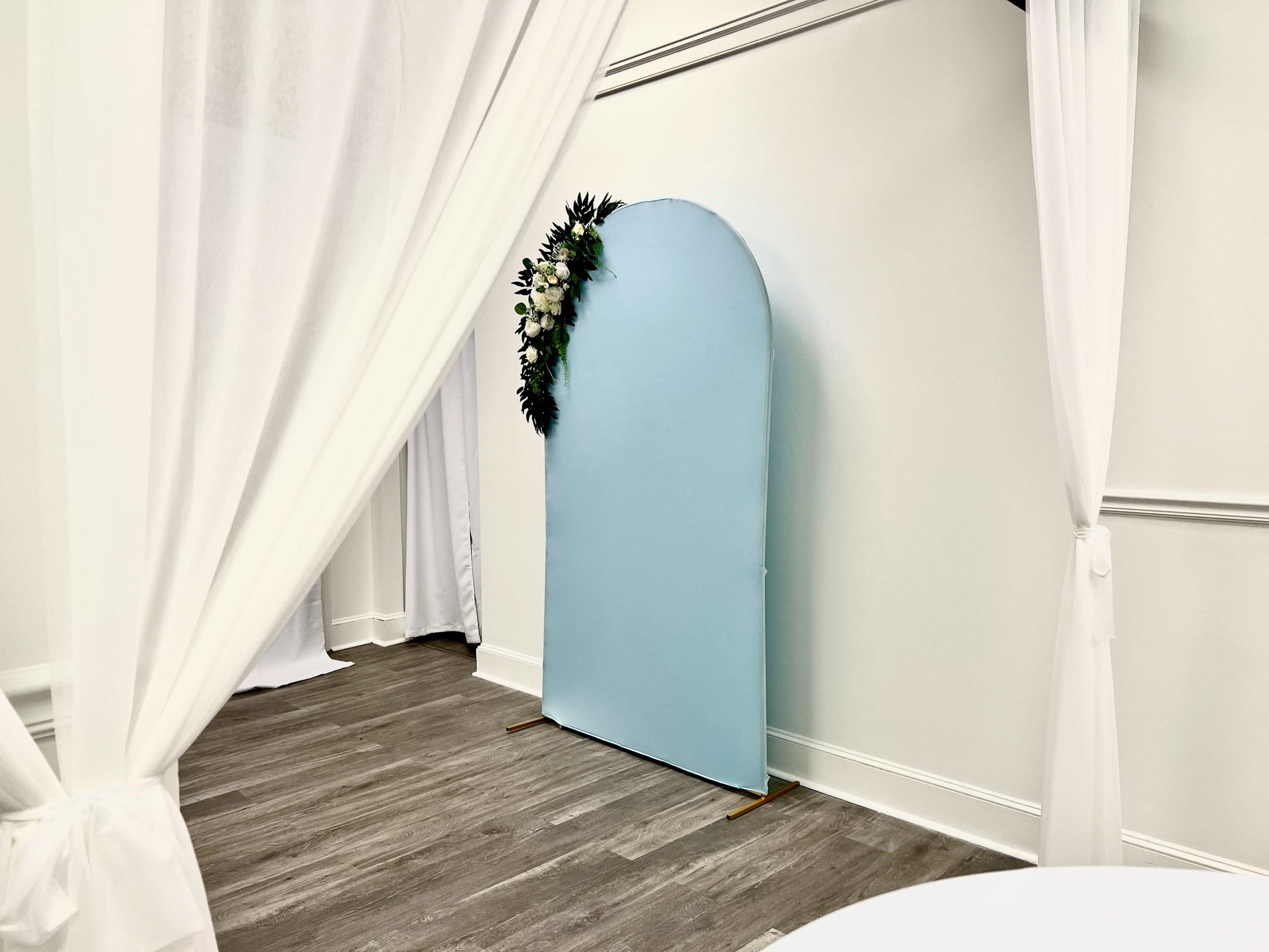 A light blue photo backdrop adorned with greenery is positioned against a white wall in a room with draped white fabric.