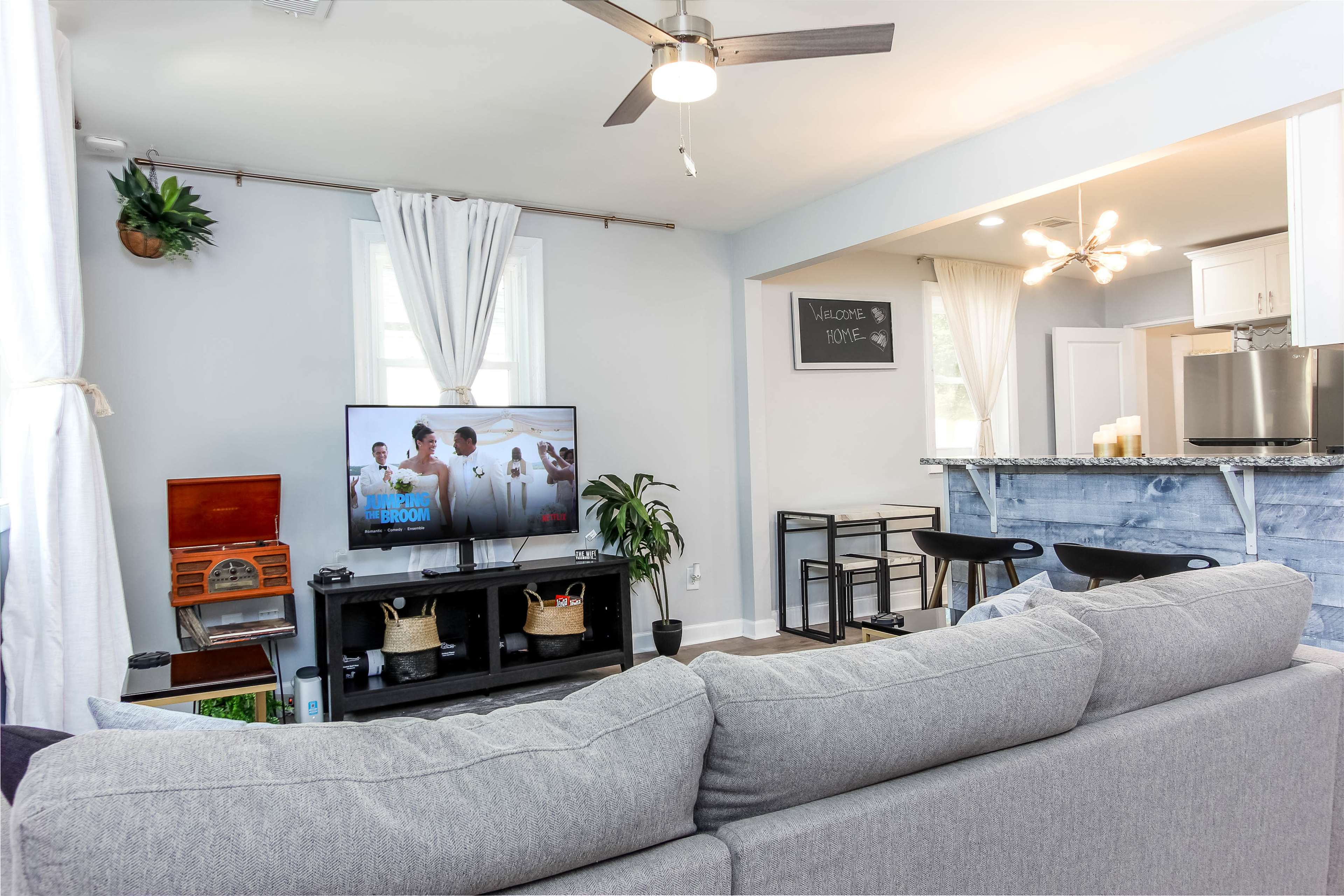 A modern living room features a gray couch, a television displaying a group of people, and a bar area with high stools in the background.