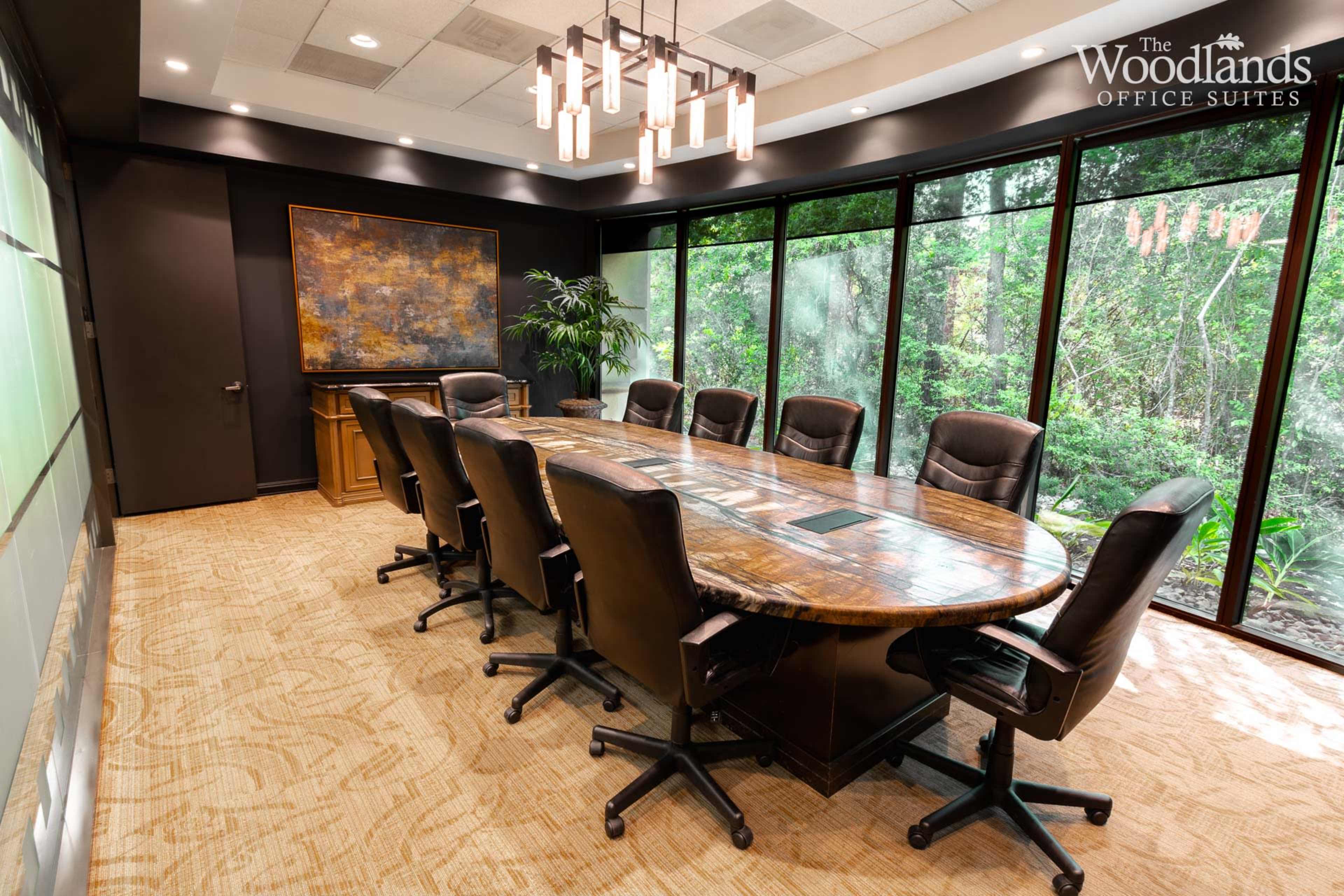 A conference room features a large round wooden table surrounded by executive chairs, with large windows overlooking a green landscape.