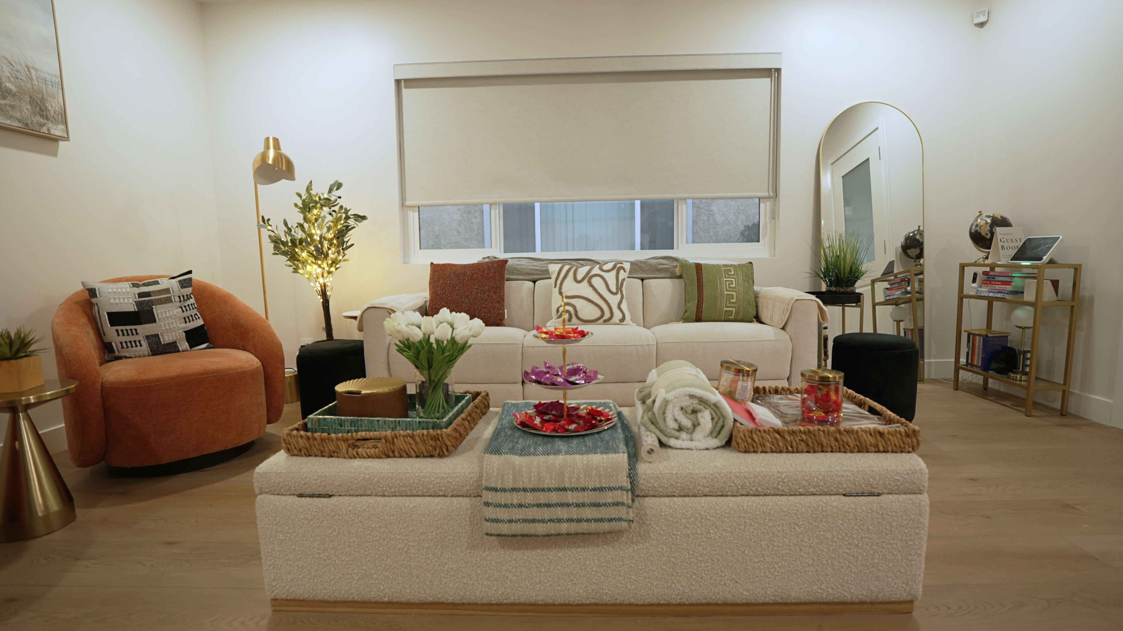 A cozy living room features a light-colored sofa adorned with decorative pillows, surrounded by a stylish armchair, potted plants, and a coffee table arrangement of vibrant snacks and flowers.