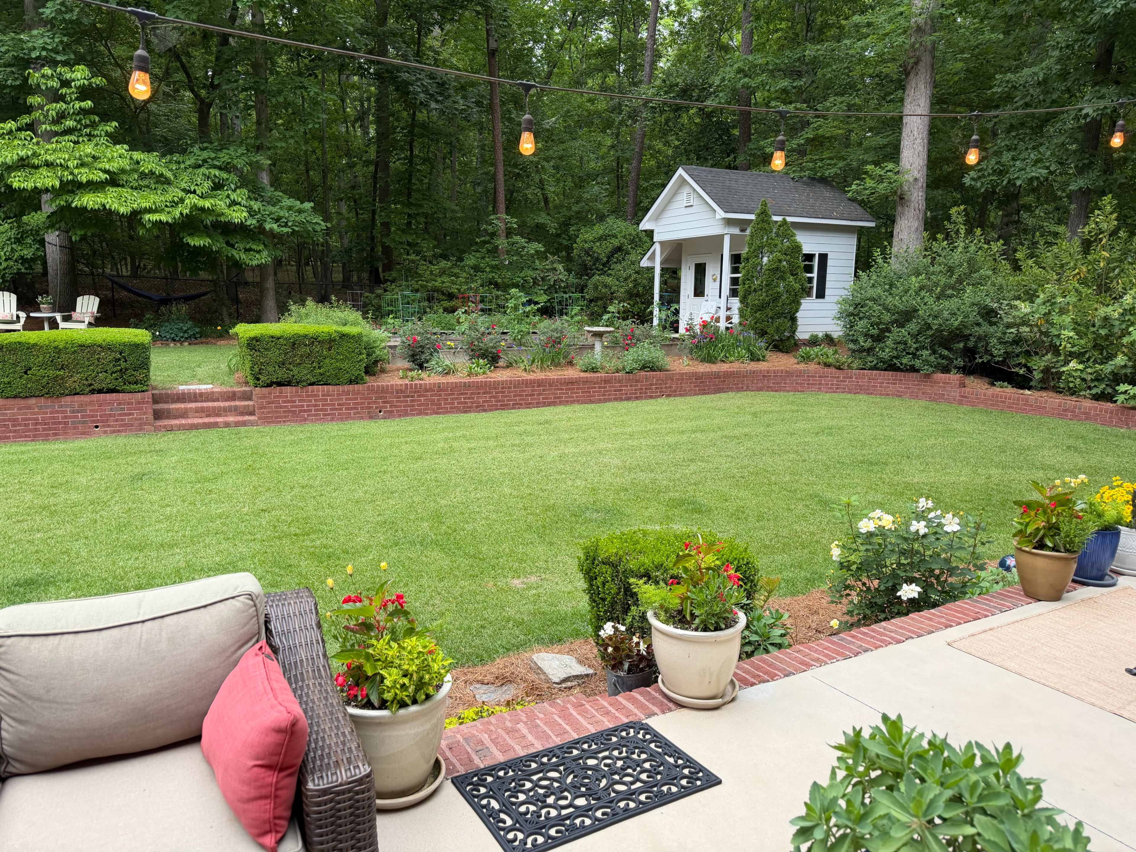 The image shows a neatly maintained backyard with green grass, flower beds, a small white shed, and outdoor seating.
