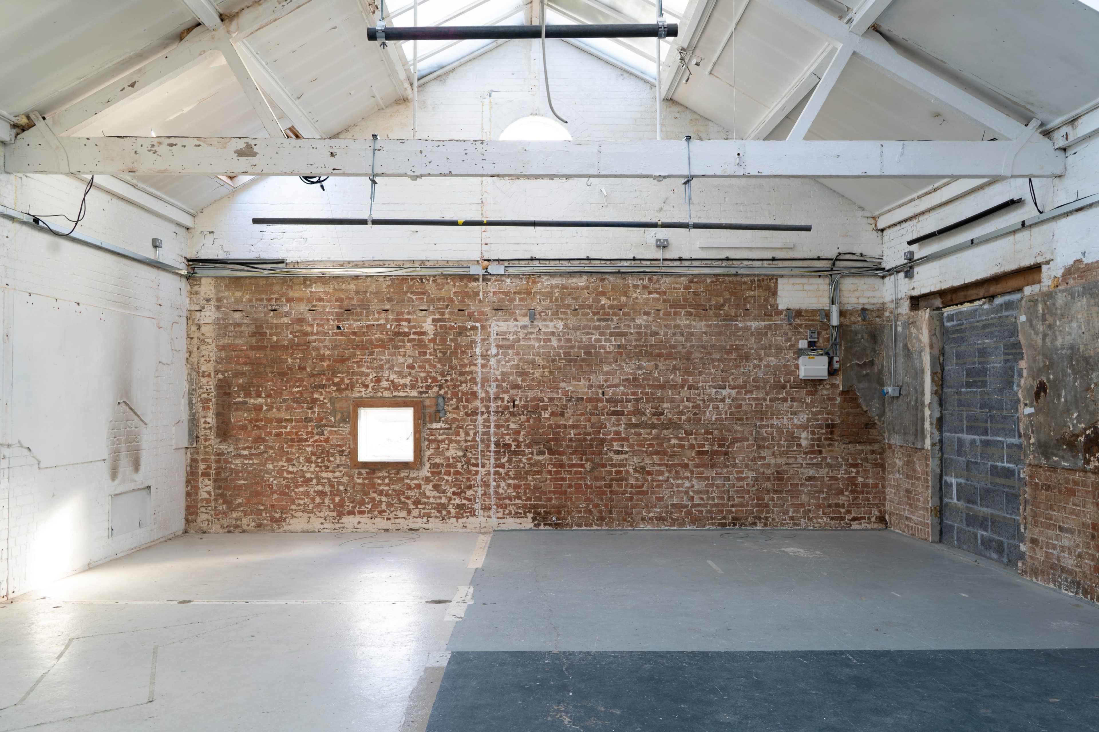 The image shows an empty warehouse interior with exposed brick walls, a high ceiling, and a concrete floor.
