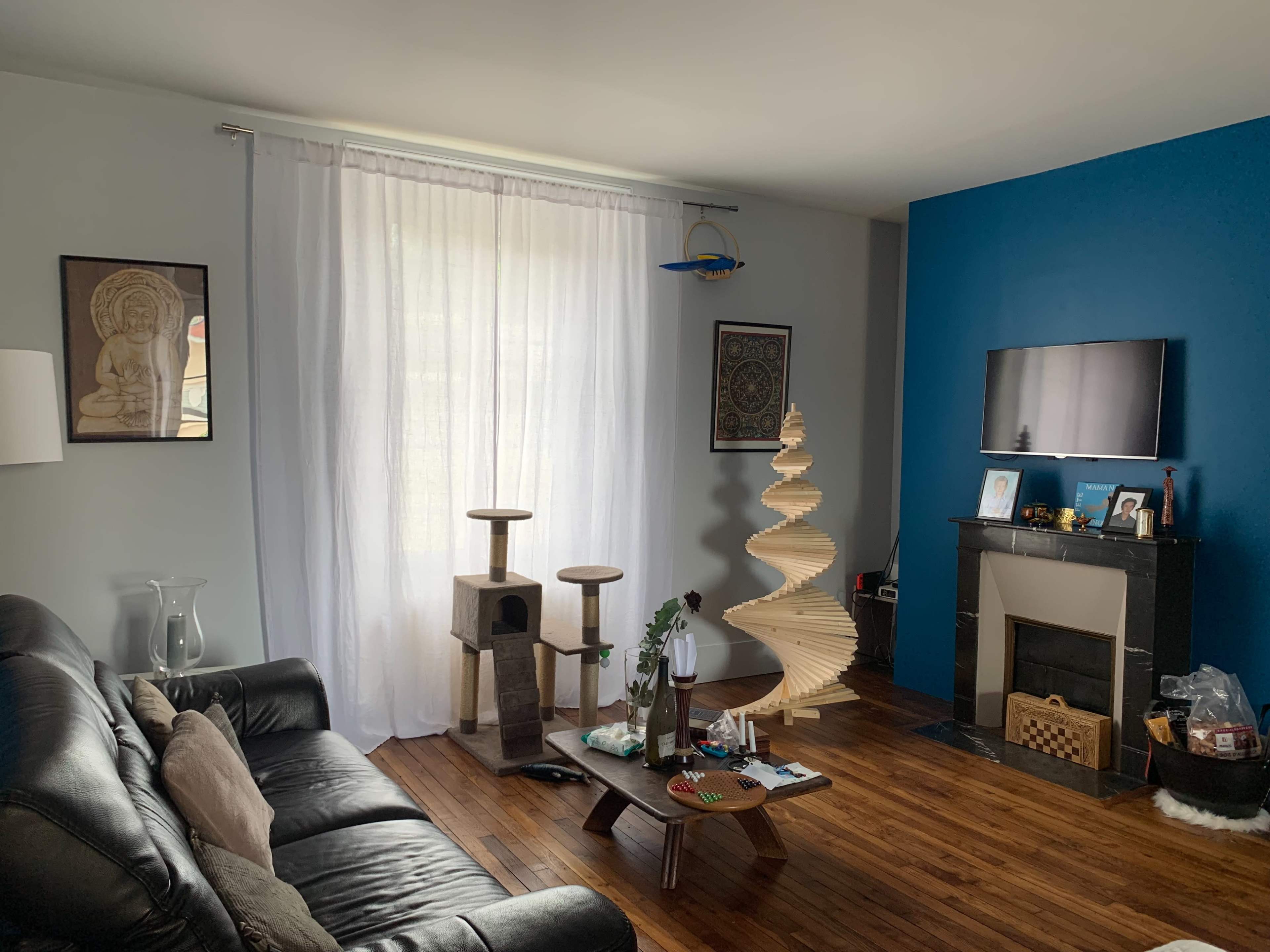 The image shows a living room with a black leather sofa, a wooden floor, a tall decorative cat tree, a spiral bookshelf, and a wall-mounted TV against a blue feature wall.