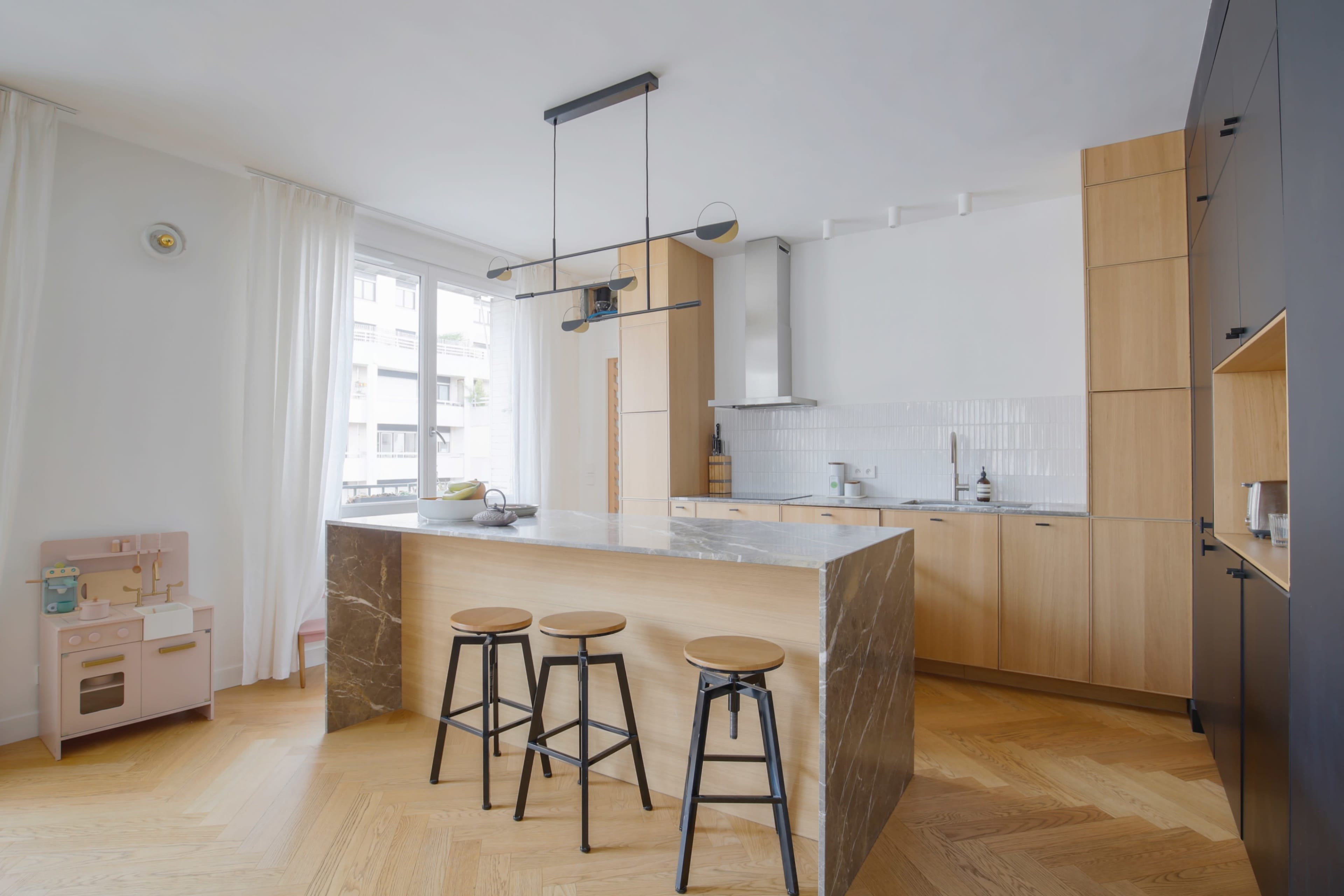 A modern kitchen featuring a large marble island with three black stools, wooden cabinetry, and large windows allowing natural light.