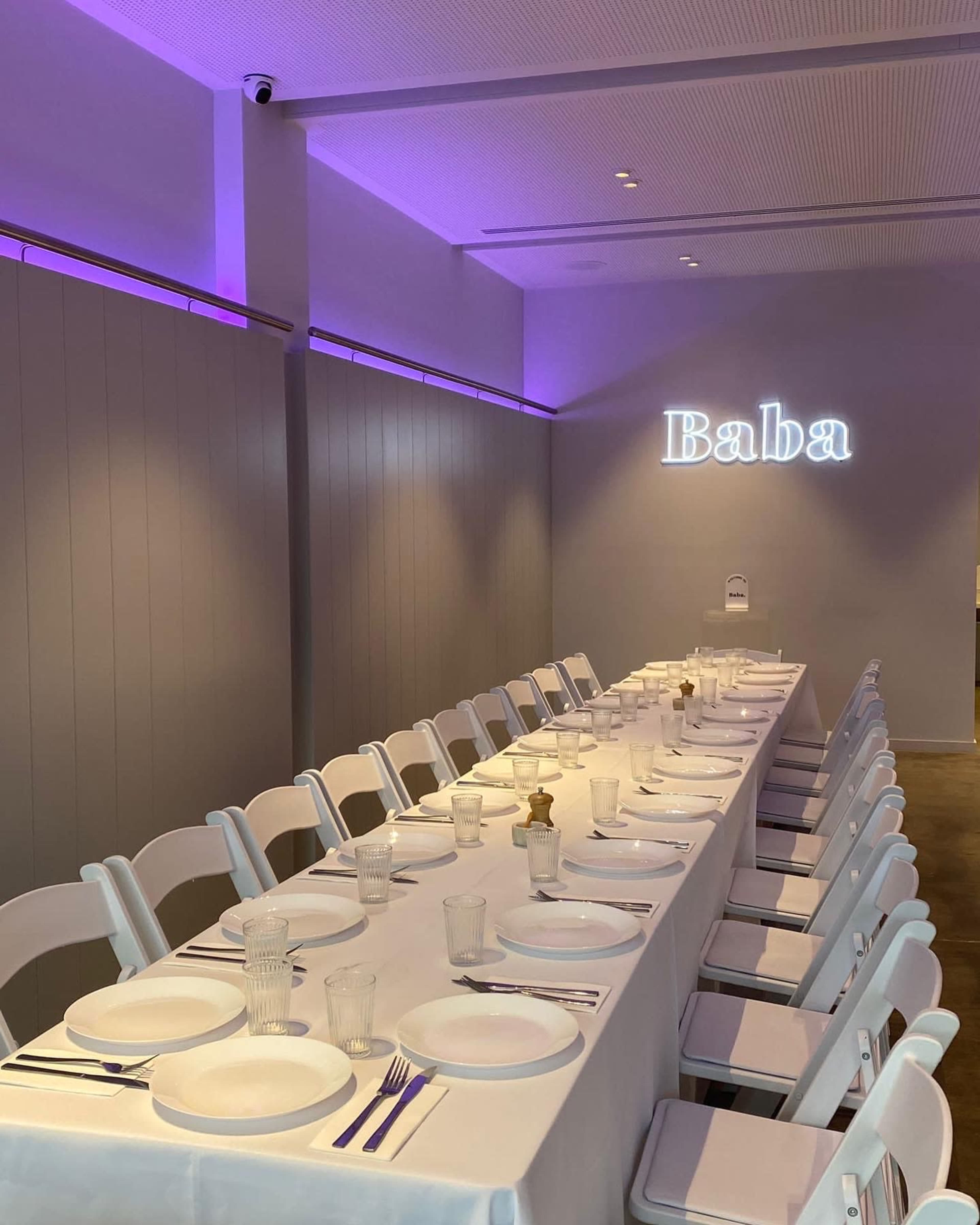 A long, neatly set dining table with white tableware and clear glasses is illuminated by purple lighting, with the word "Baba" displayed on the wall.